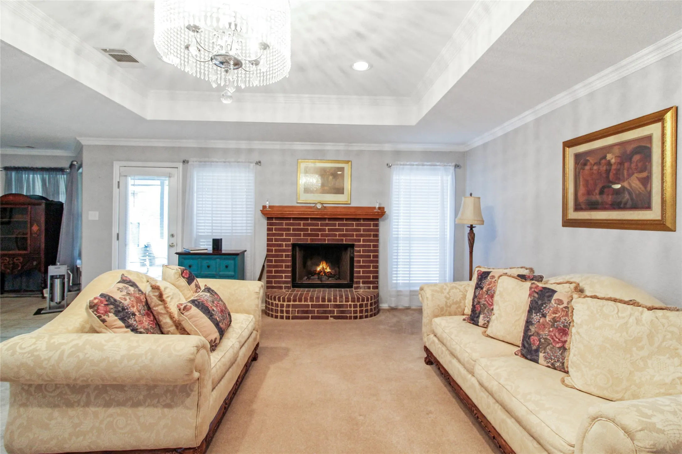 Living area featuring a brick fireplace with wood mantel, a coffered ceiling with a crystal chandelier, and two large windows with blinds
