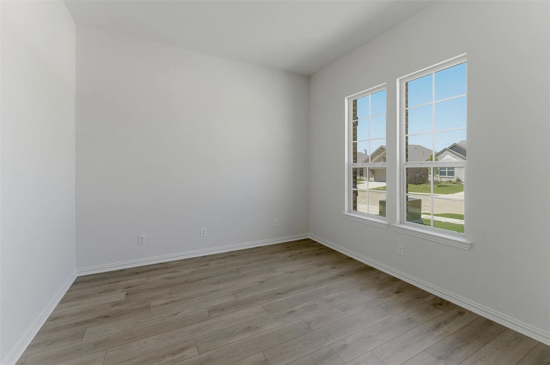 Spacious room featuring wood-finish flooring, two large windows with grid patterns, bright white walls, white baseboards, and multiple electrical outlets