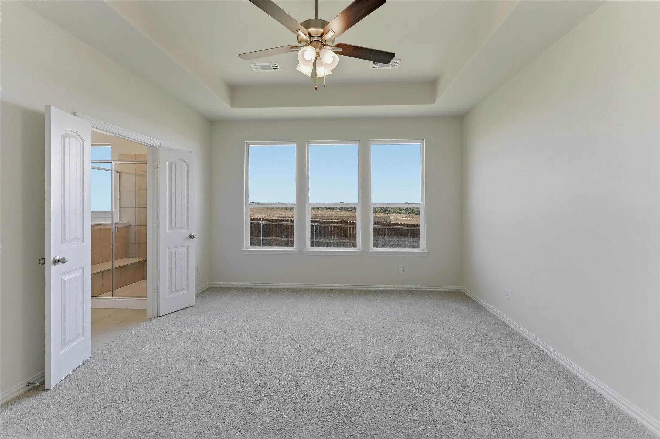 Spacious carpeted room featuring a ceiling fan with light fixture, tray ceiling, and three large windows