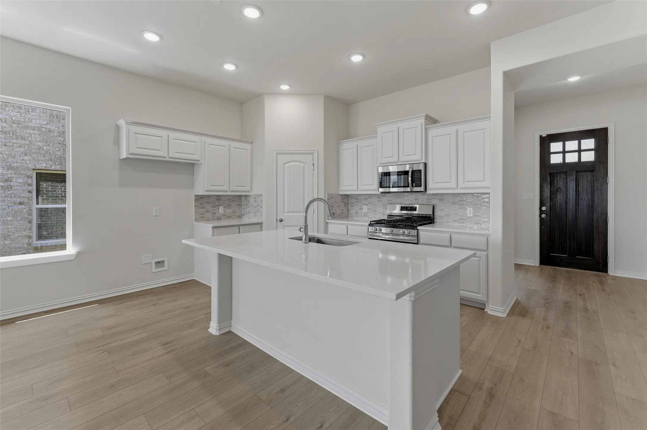 Bright kitchen featuring a central island with an undermount sink, white shaker cabinetry, stainless steel appliances, a tile backsplash, and recessed lighting