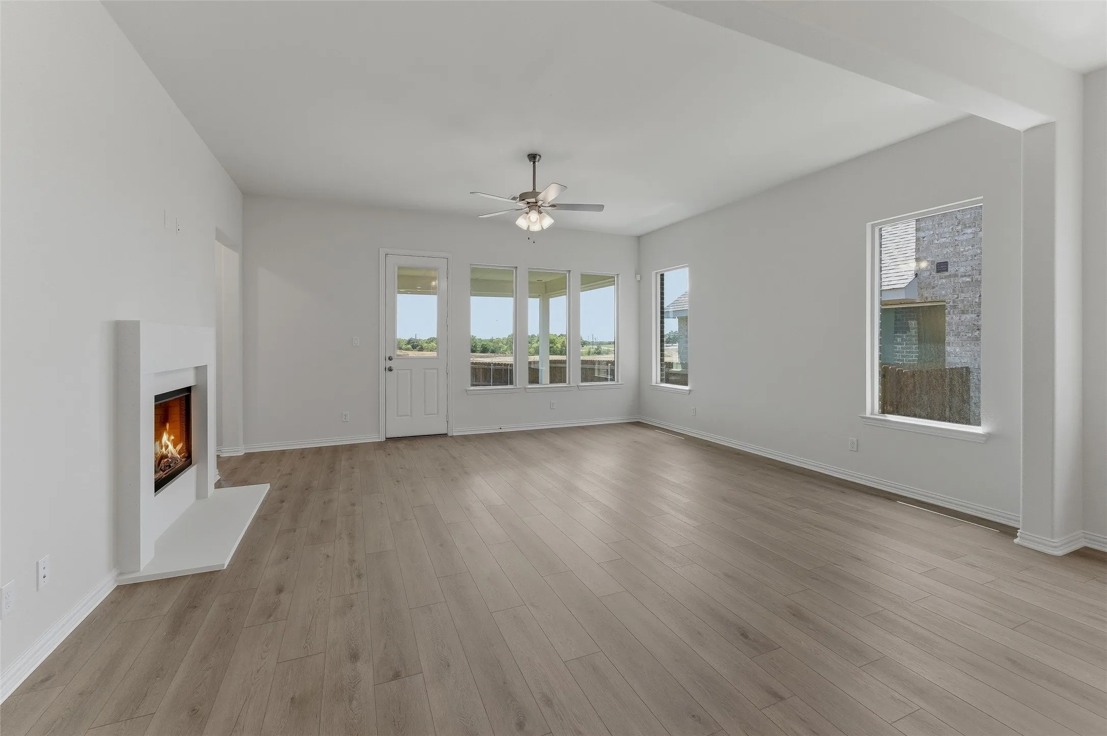 Spacious living area featuring light wood-finish flooring, a modern white fireplace, multiple windows with exterior views, and a ceiling fan