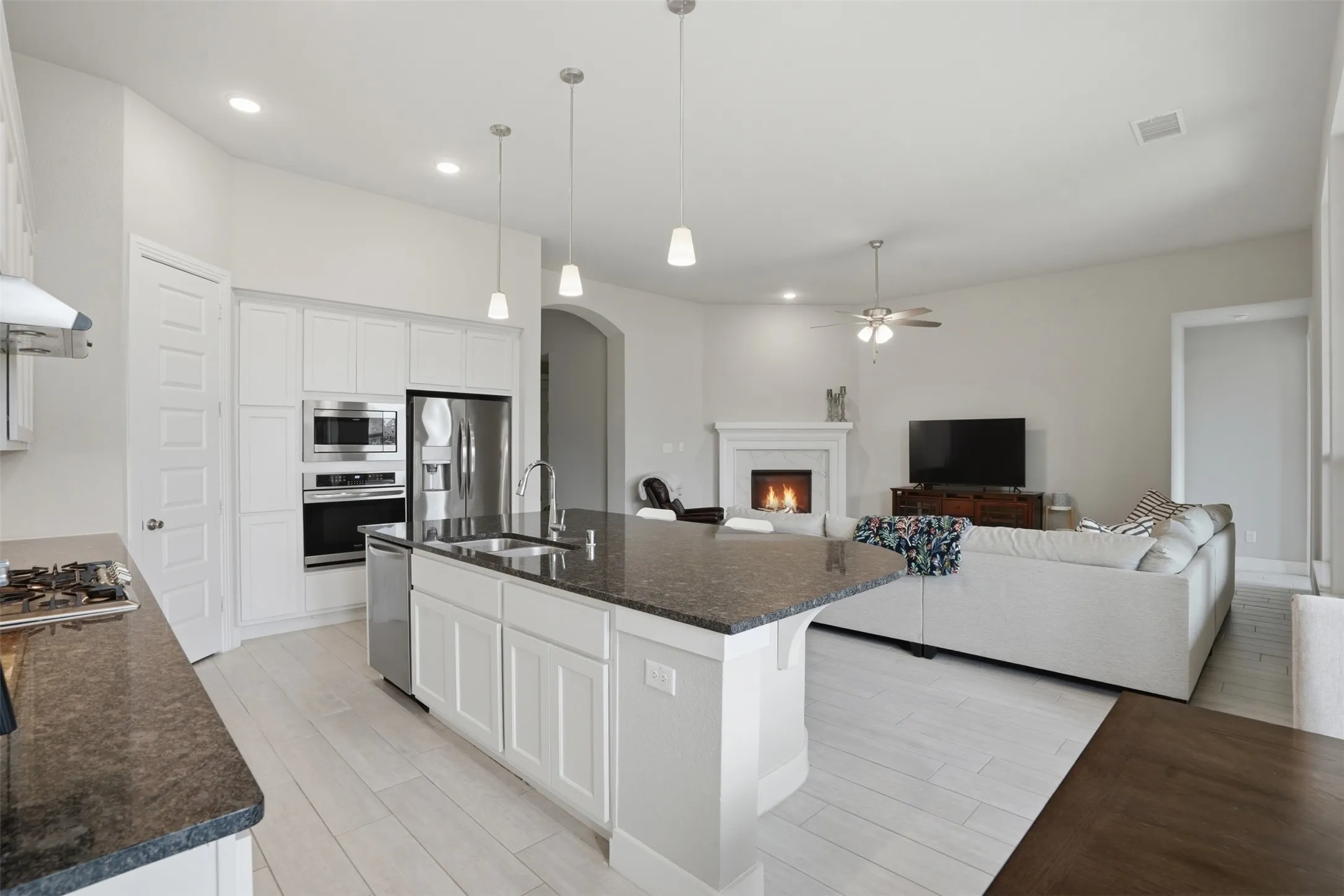 Open concept living space featuring a modern kitchen with a large island, stainless steel appliances, and light-colored flooring, opening into a living room with a fireplace and a ceiling fan