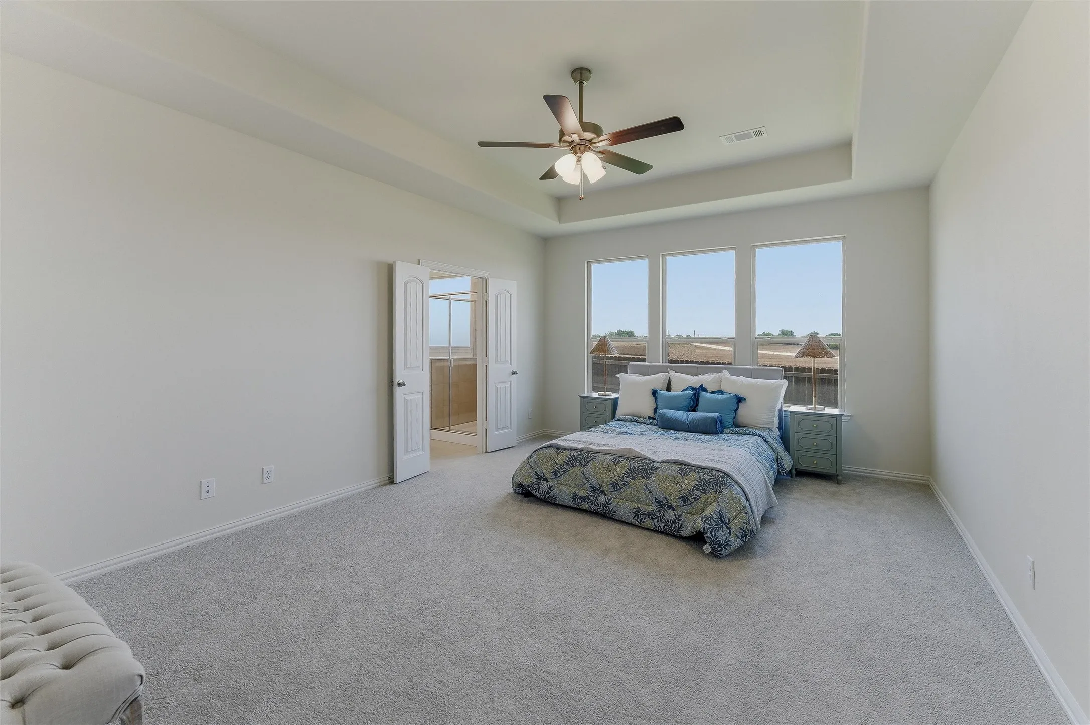 Spacious room featuring light gray carpeting, neutral wall finishes, a tray ceiling with a ceiling fan, and three large windows