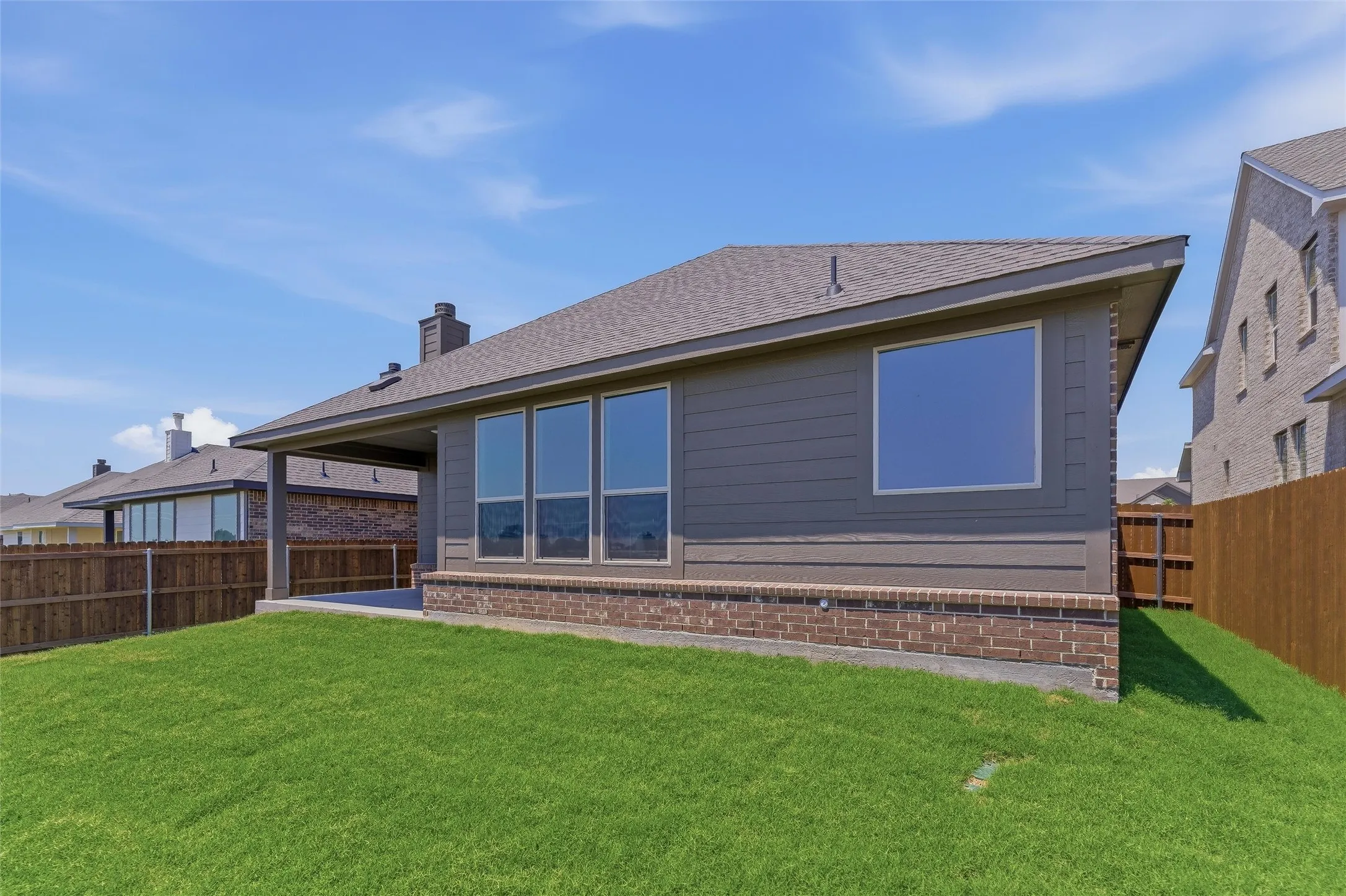 Expansive backyard featuring a manicured lawn, a covered patio, brick foundation, and multiple large windows on the rear elevation