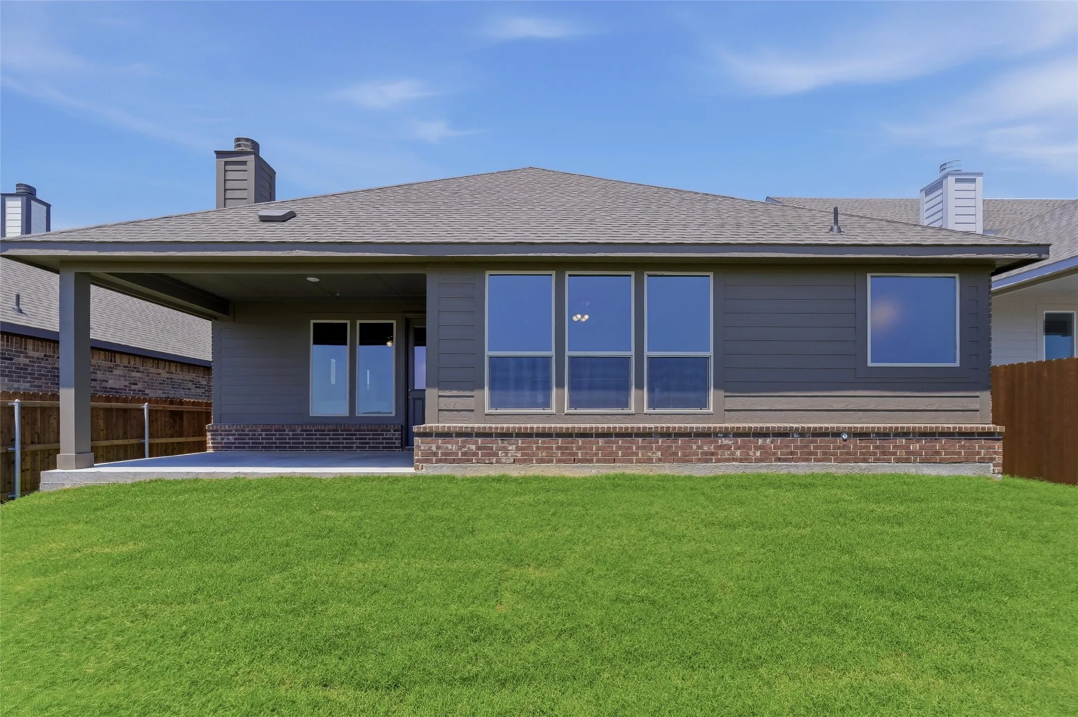 Expansive green lawn with a covered patio, large windows, brick detailing, and a shingle roof