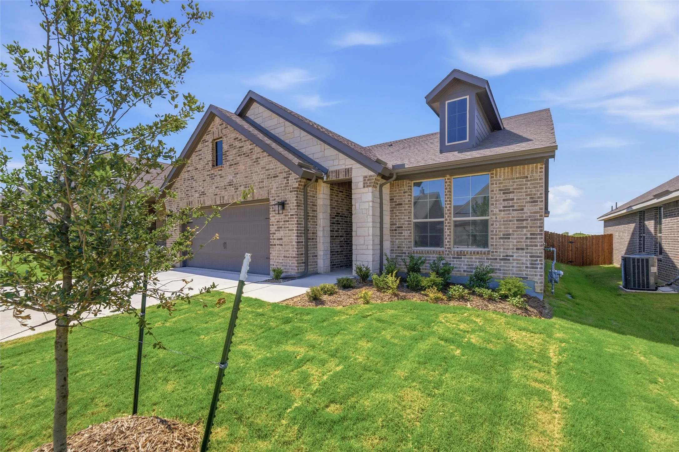 Brick and stone facade residence featuring an attached garage, a dormer window, and mature landscaping