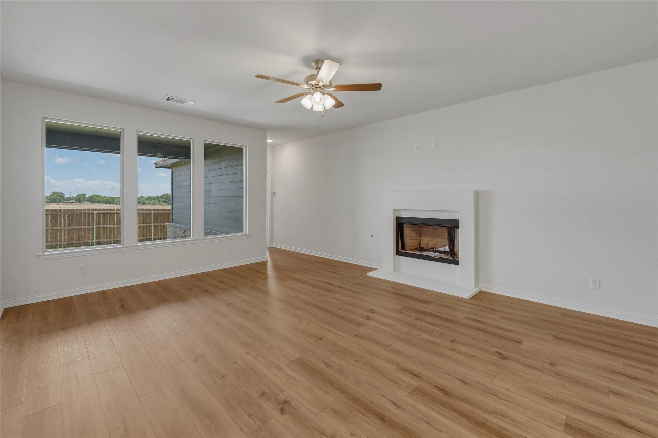 Spacious room featuring wood-finish flooring, a contemporary fireplace with a white surround, and three large windows
