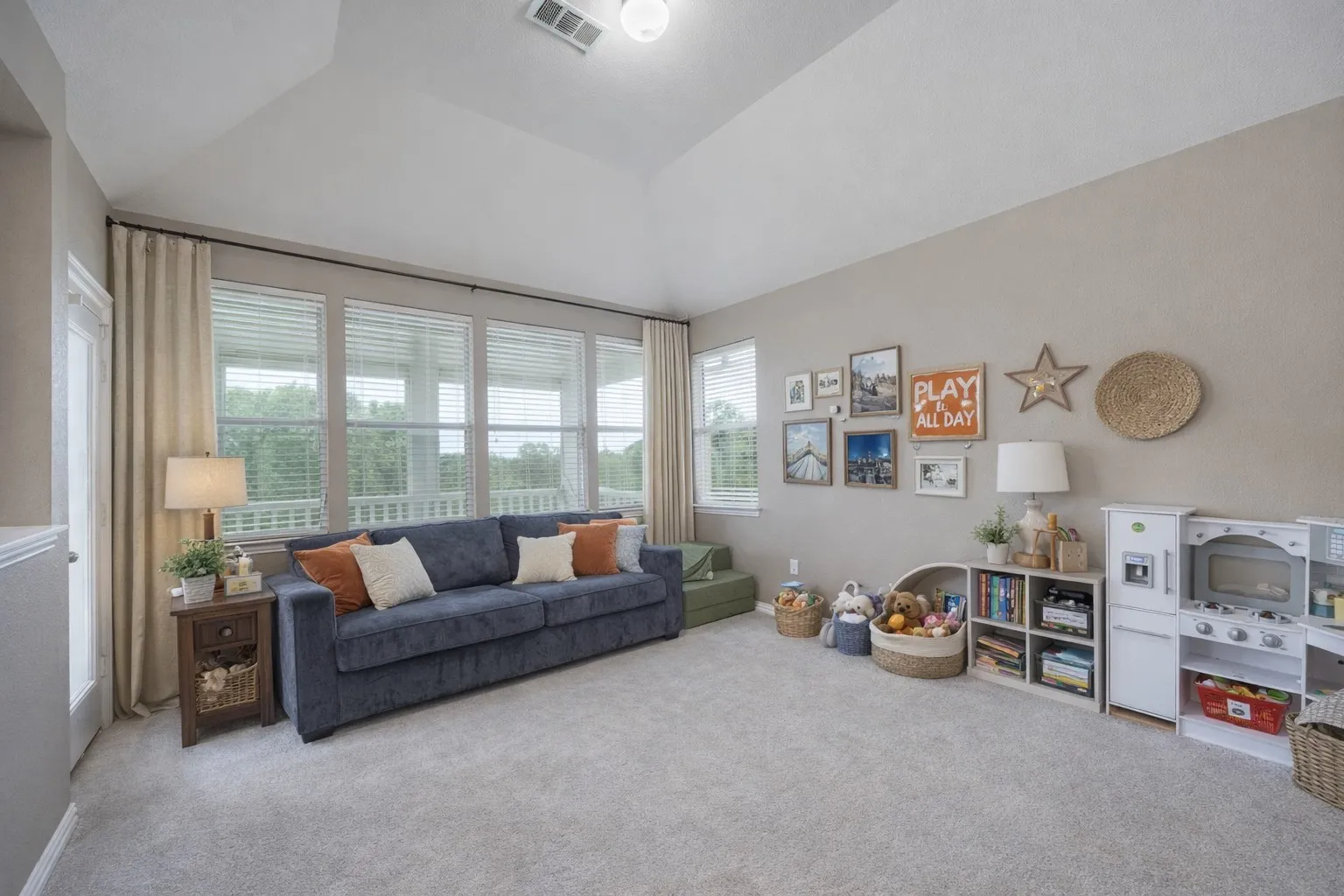 This room features light-colored carpeting, a vaulted ceiling, and a wall of windows fitted with blinds and drapes, offering views of the outdoors