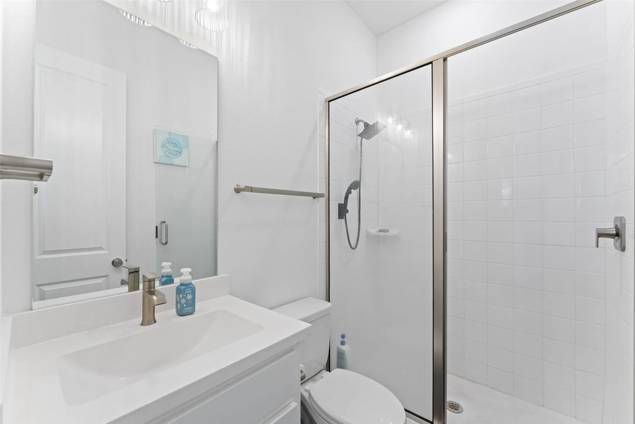 Bathroom featuring a vanity with an integrated sink, a large mirror, a toilet, and a glass-enclosed shower with a tiled surround