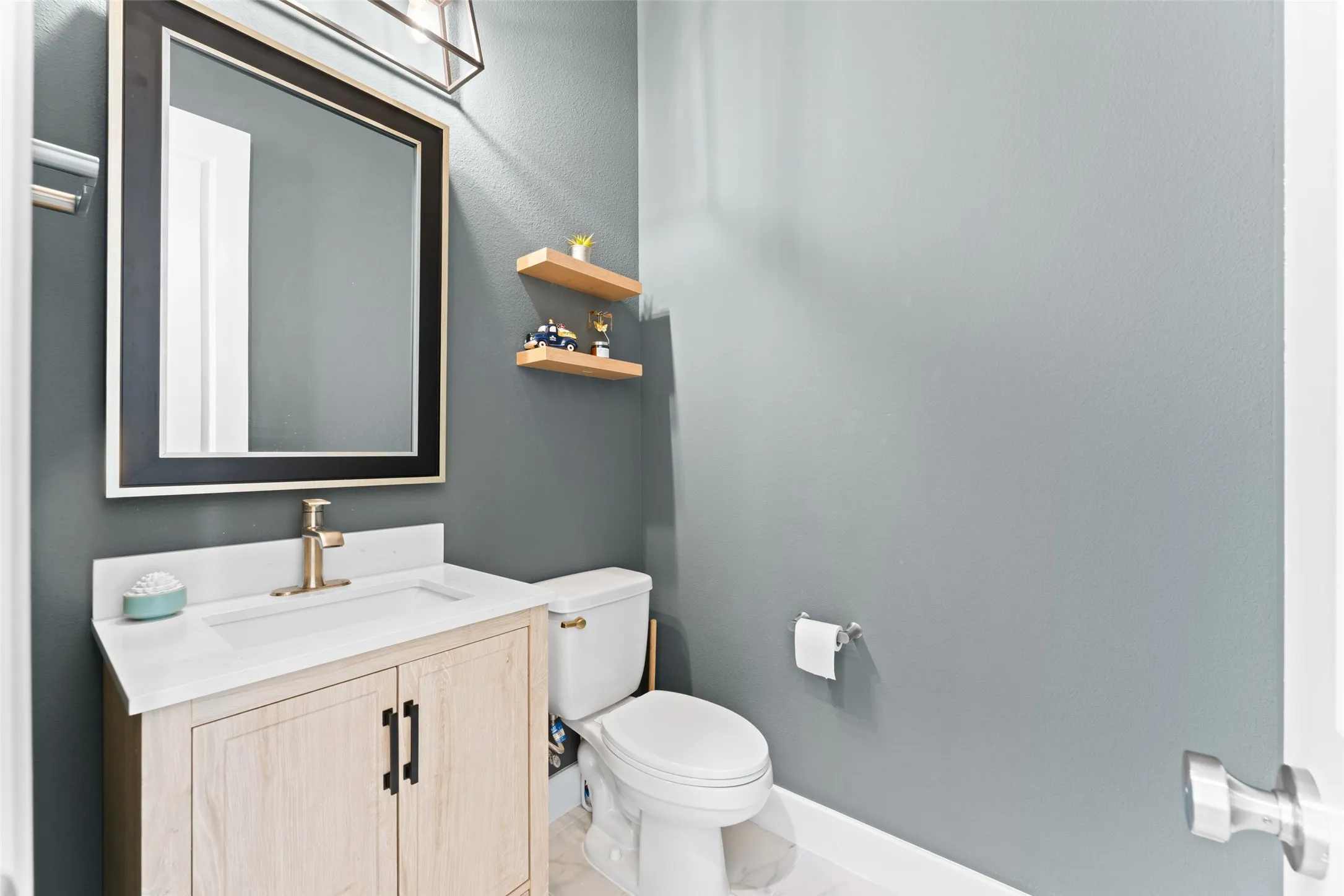 A well-appointed powder room featuring a light wood vanity with a white countertop and a gold-toned faucet