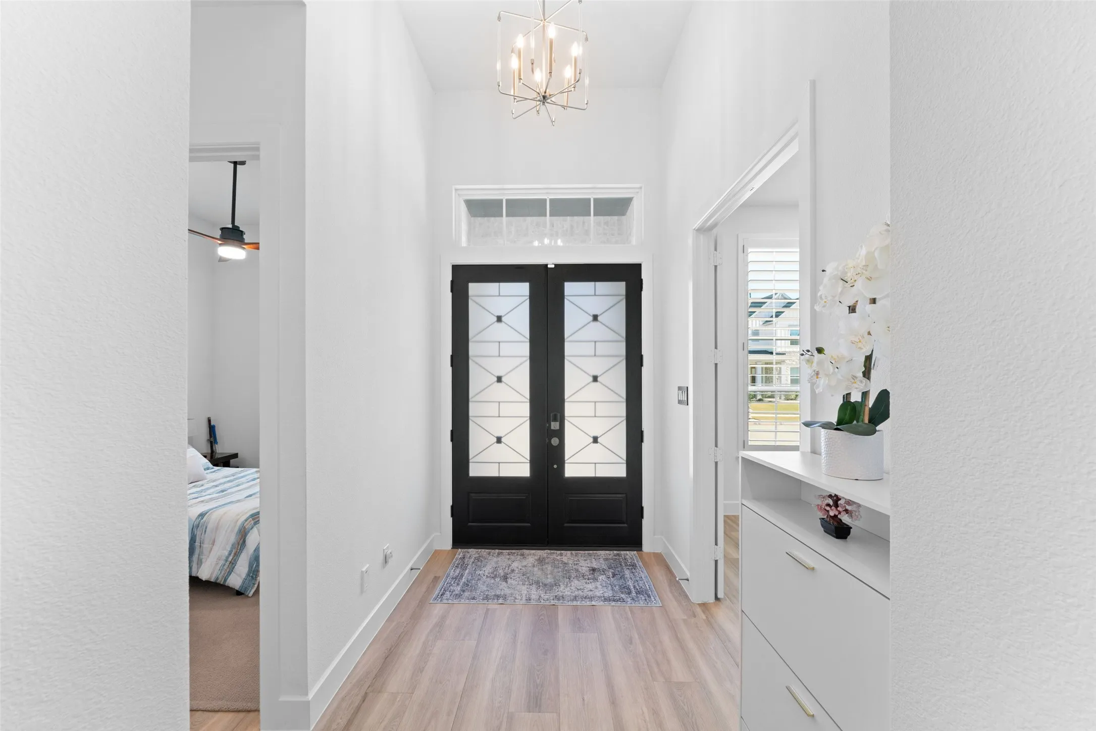 The entryway features double black doors with decorative glass panels, light wood flooring, and a modern chandelier