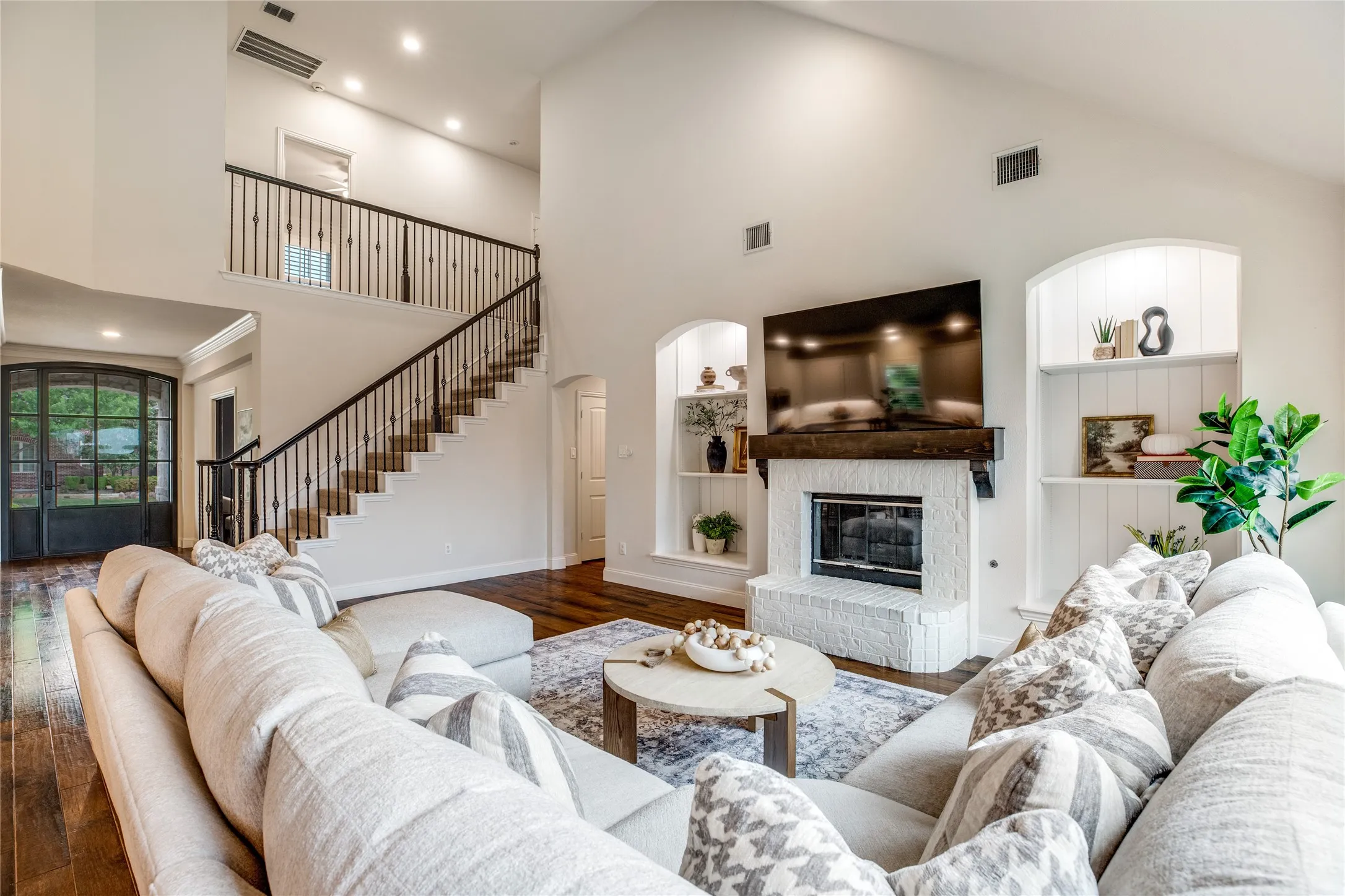 Spacious living area featuring high vaulted ceilings, rich hardwood flooring, a white brick fireplace with a wood mantle, and built-in shelving units