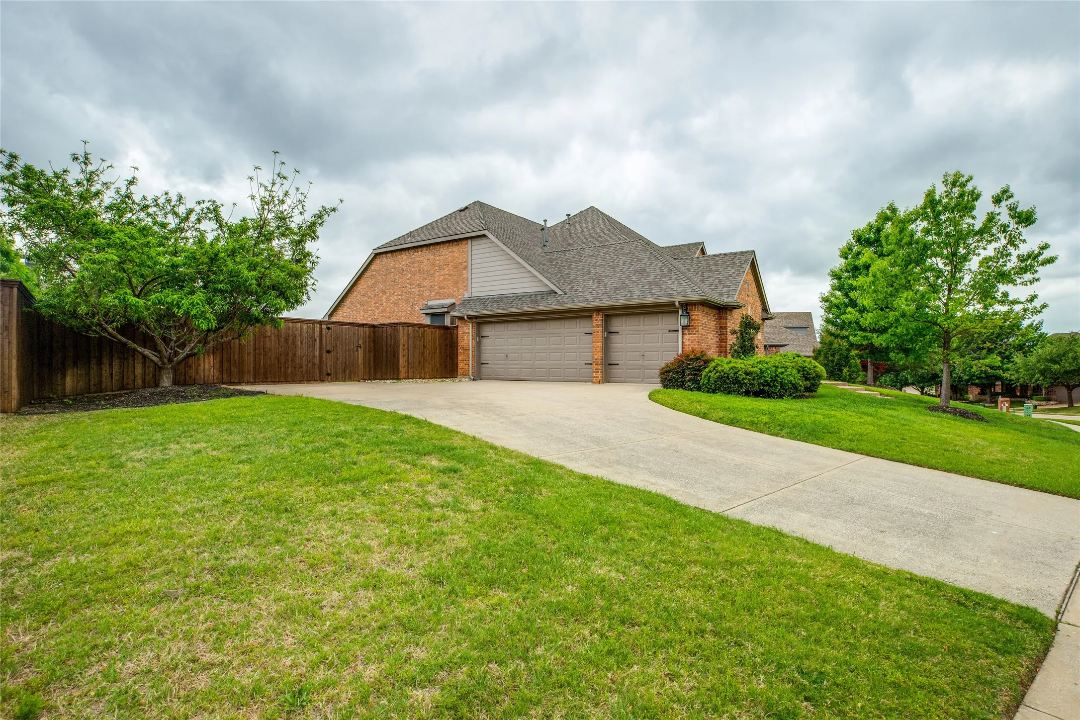 The property features a concrete driveway leading to a two-car garage, brick exterior, and a well-maintained lawn with mature trees and shrubs