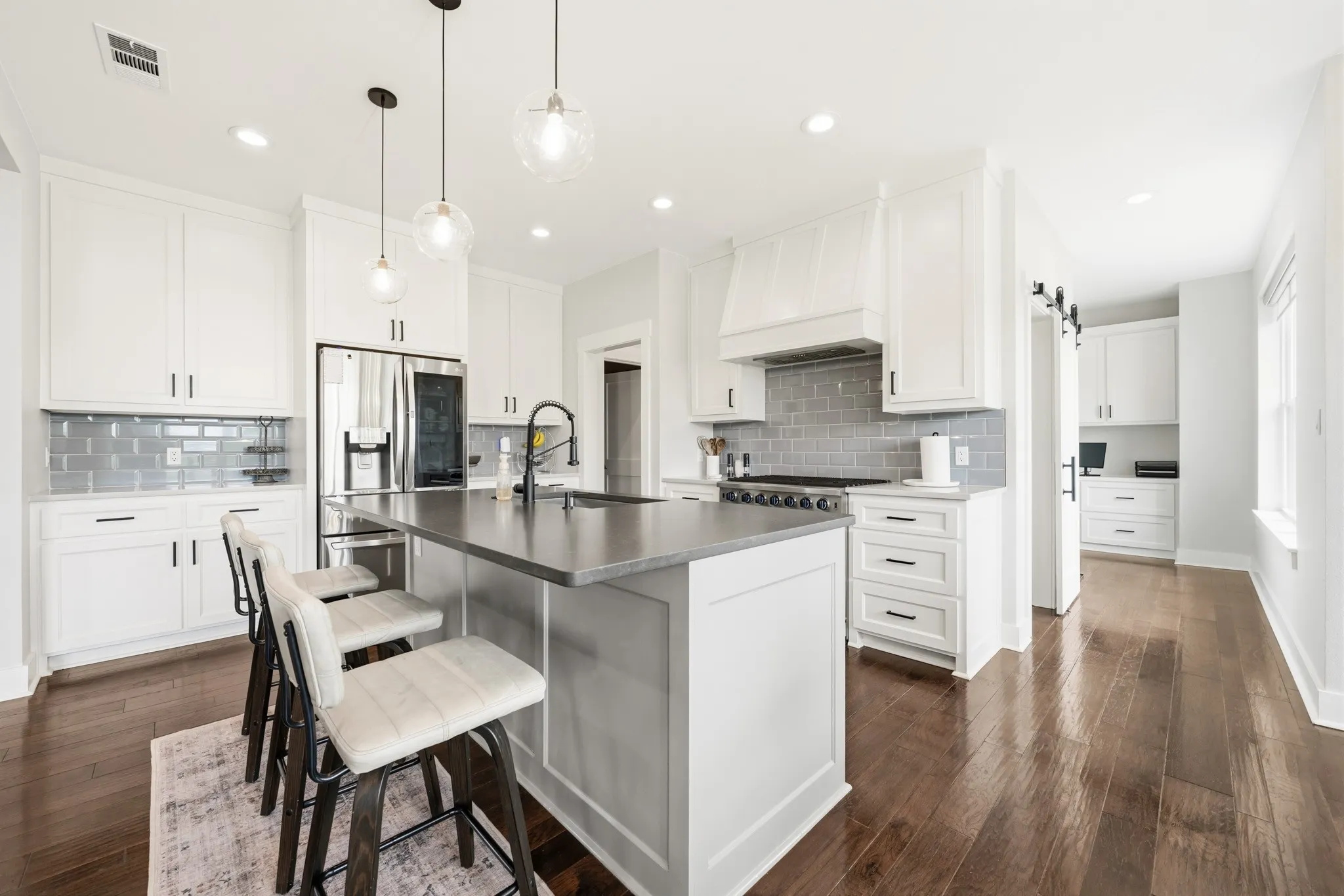 The kitchen features an island with a dark countertop and pendant lighting, white cabinetry, a grey subway tile backsplash, and dark wood flooring