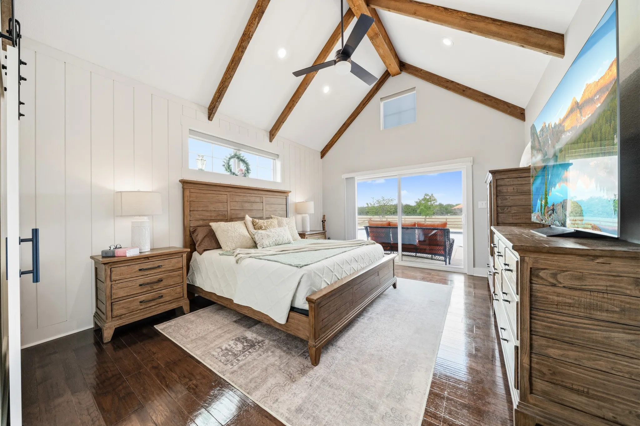 The bedroom features high vaulted ceilings with exposed wood beams, rich hardwood flooring, and a sliding glass door leading to an outdoor patio with seating