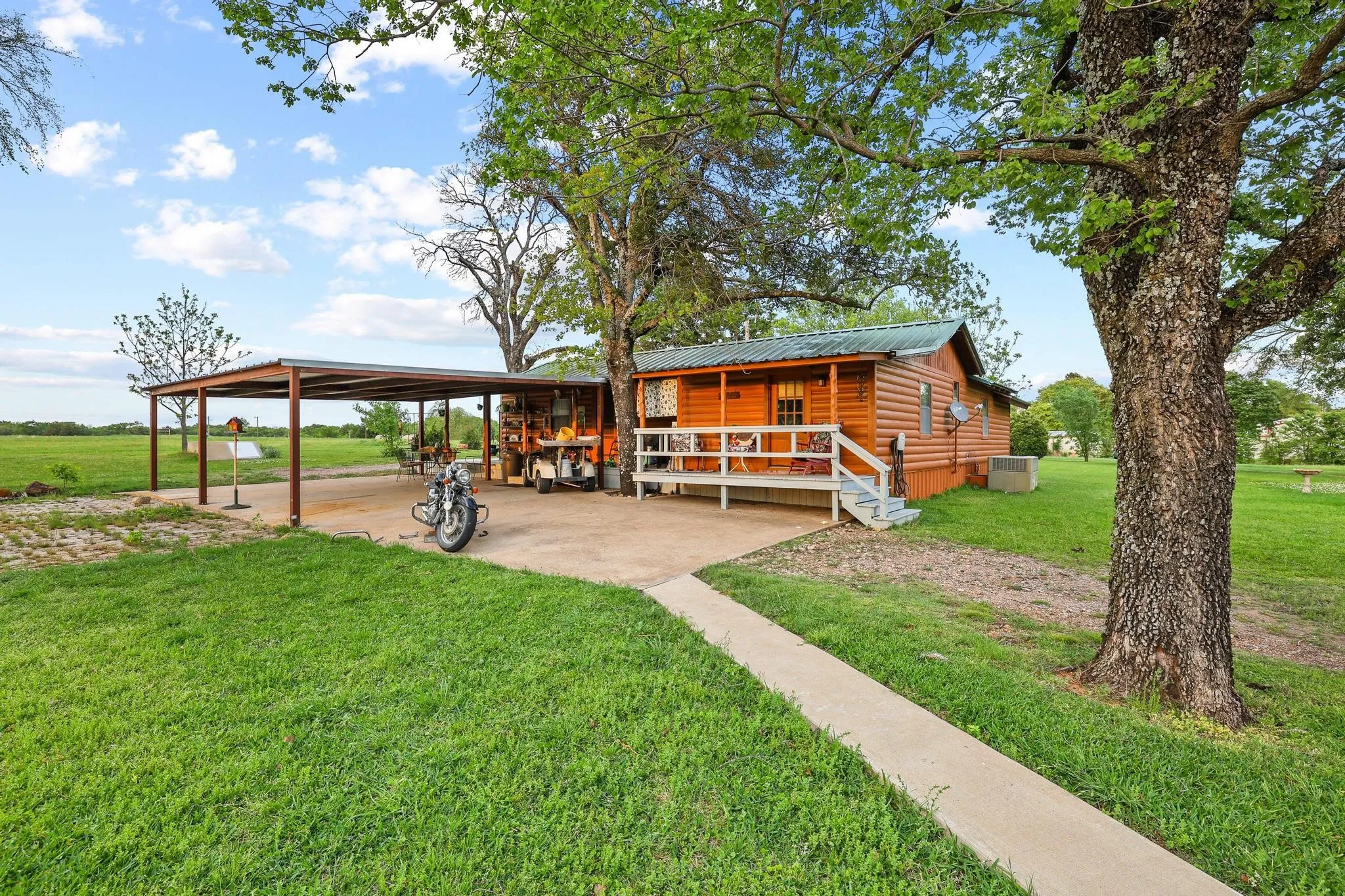 This property features a rustic cabin with a covered porch, a separate metal carport, and a paved walkway leading to the entrance