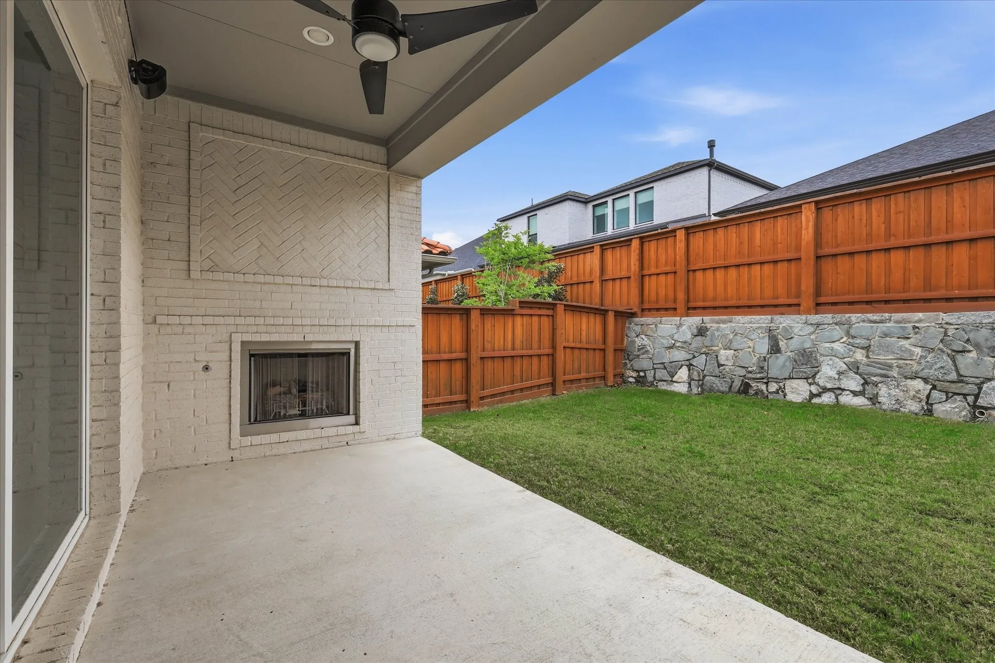 The property features a covered patio with a built-in fireplace and a ceiling fan, overlooking a private yard with a lawn, a stone retaining wall, and a wooden fence