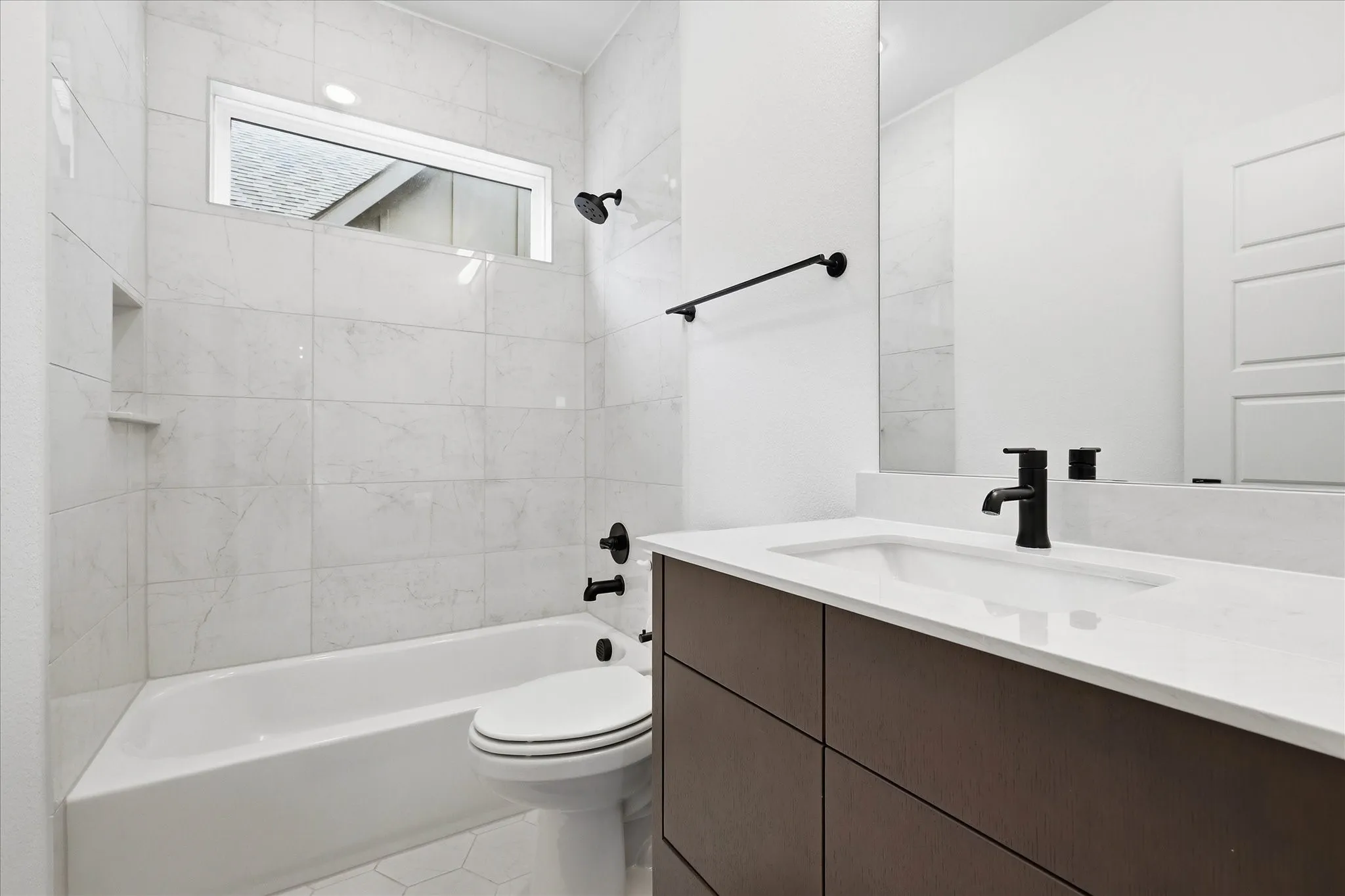 The bathroom features a bathtub with a tiled shower surround, a window above the shower, and a vanity with a white countertop and dark base