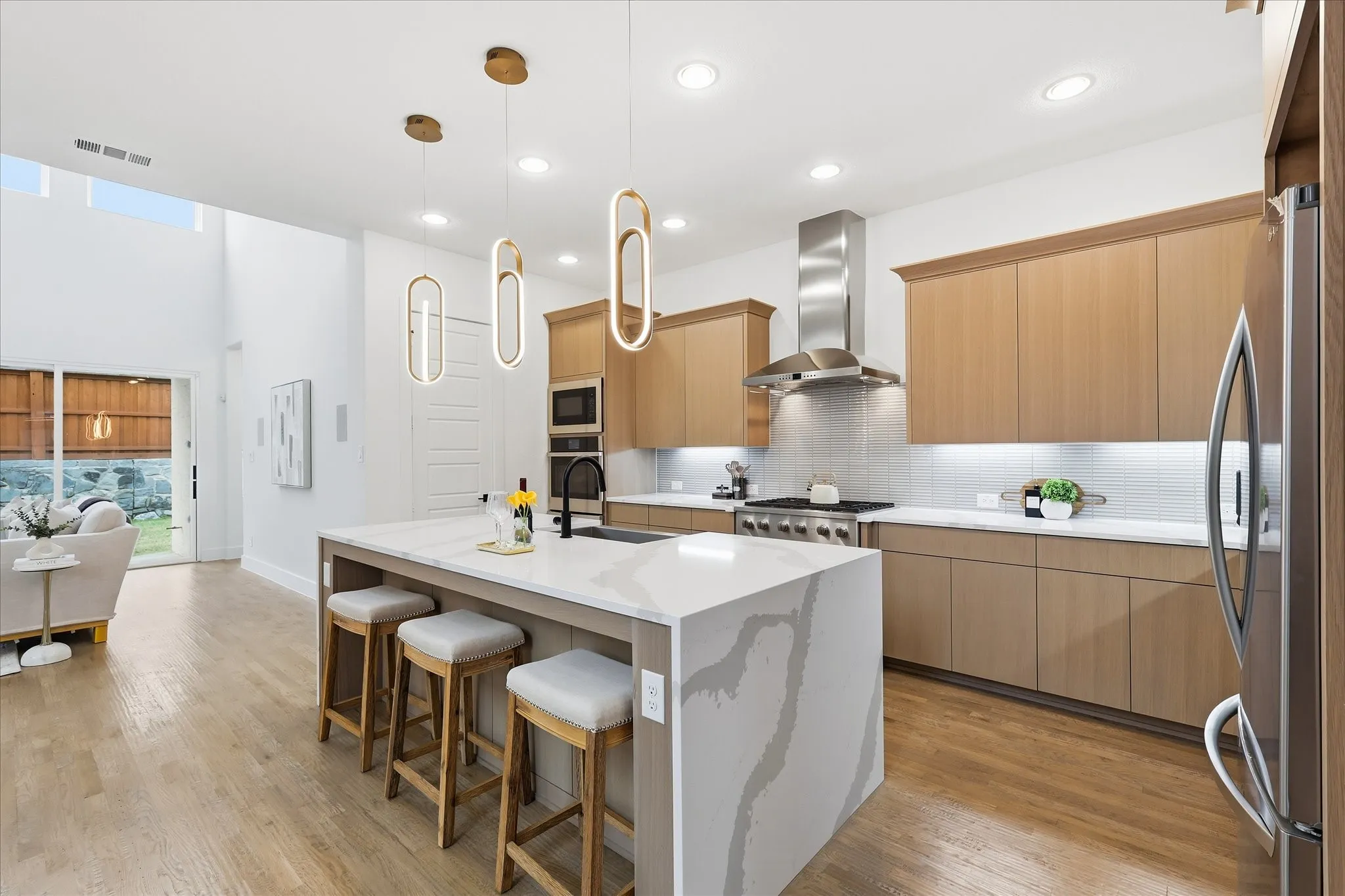 The kitchen features a large island with a quartz countertop and seating, light wood cabinetry, and stainless steel appliances including a refrigerator, oven, and range hood