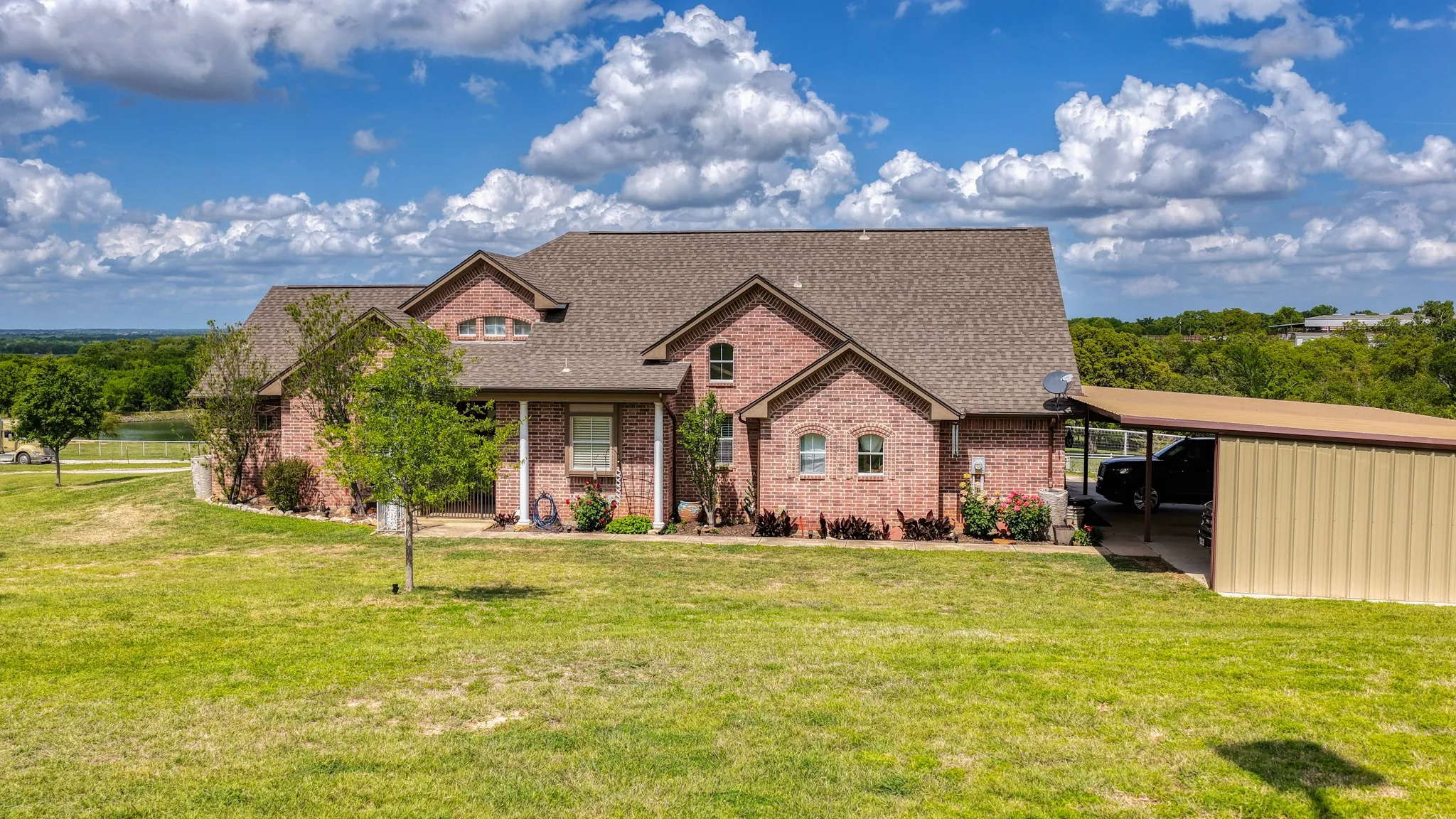 The property features a brick exterior, a substantial shingled roof, a carport, and a vast lawn with mature trees