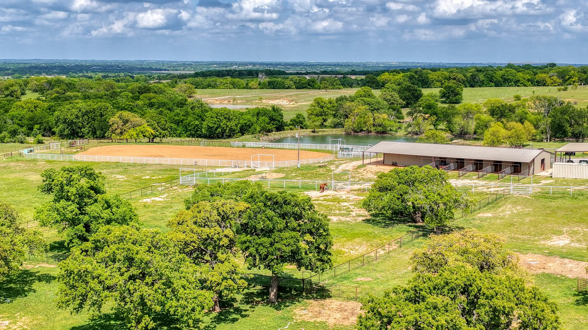 Expansive property featuring a stable building, a riding arena, and a pond, all surrounded by green pastures and mature trees