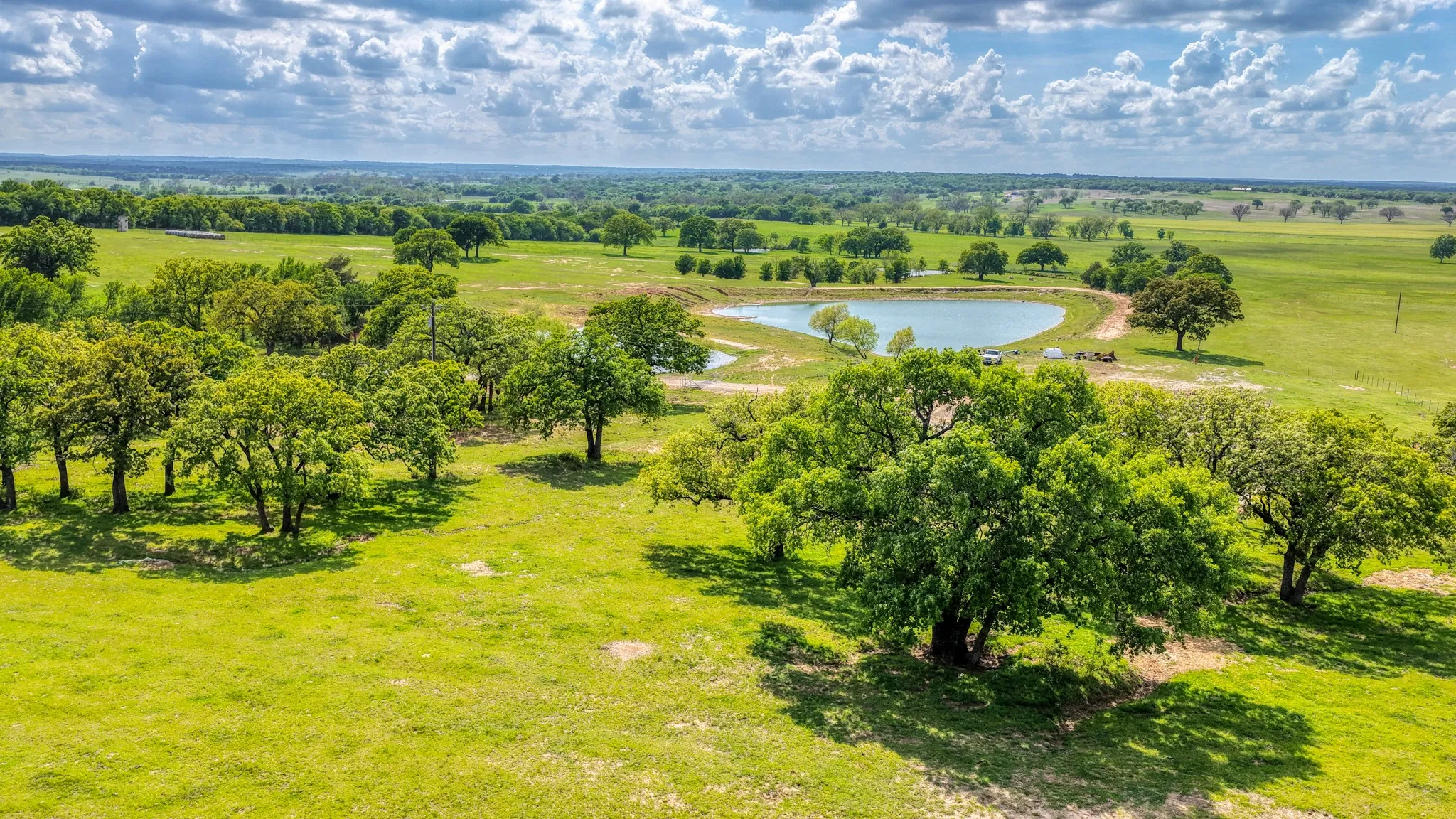 Expansive property featuring a tranquil pond, scattered mature trees, and vast green pastures under a partly cloudy sky