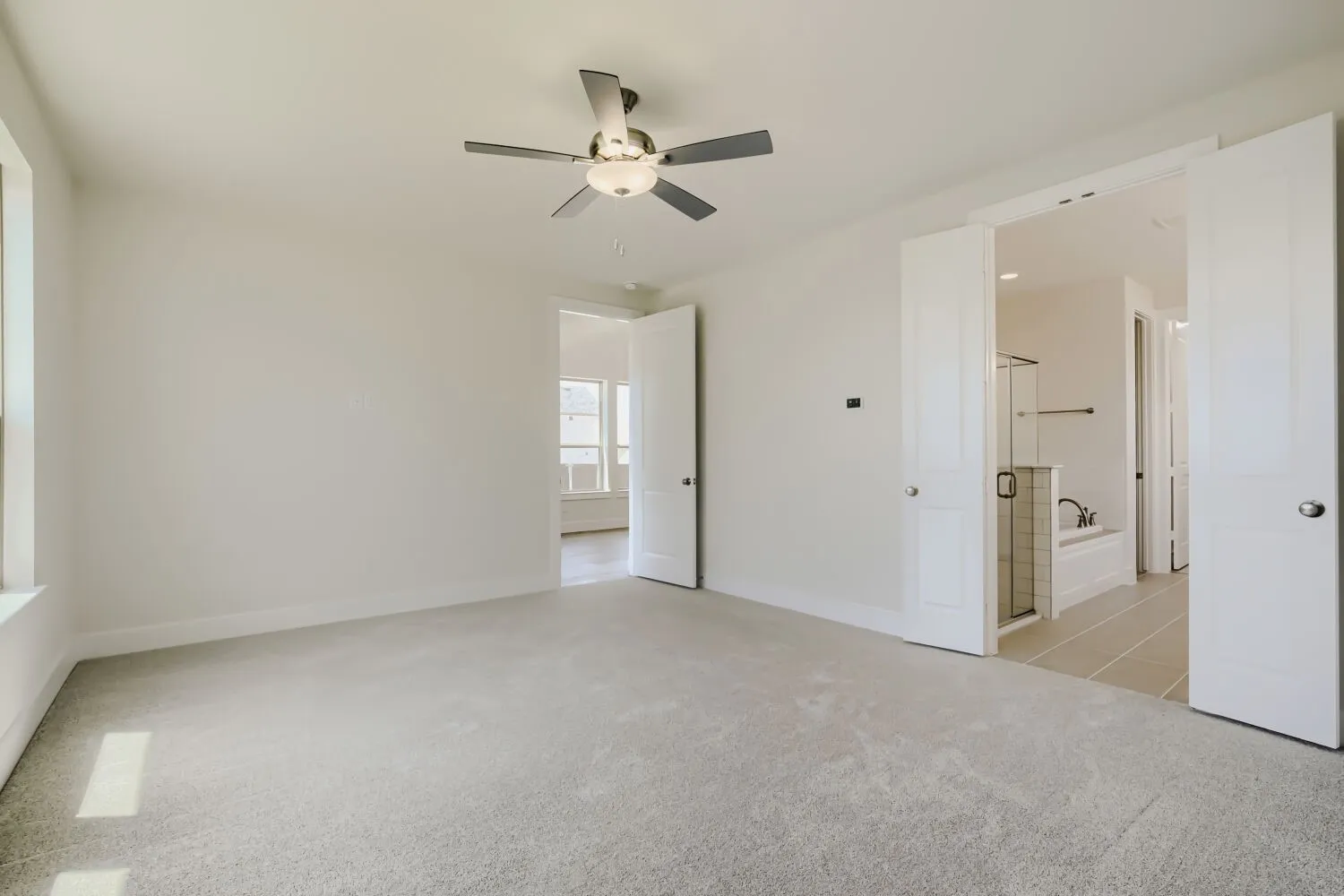 Carpeted room featuring a ceiling fan with integrated lighting, a window, and two paneled doors opening to a bathroom with a glass-enclosed shower and a bathtub