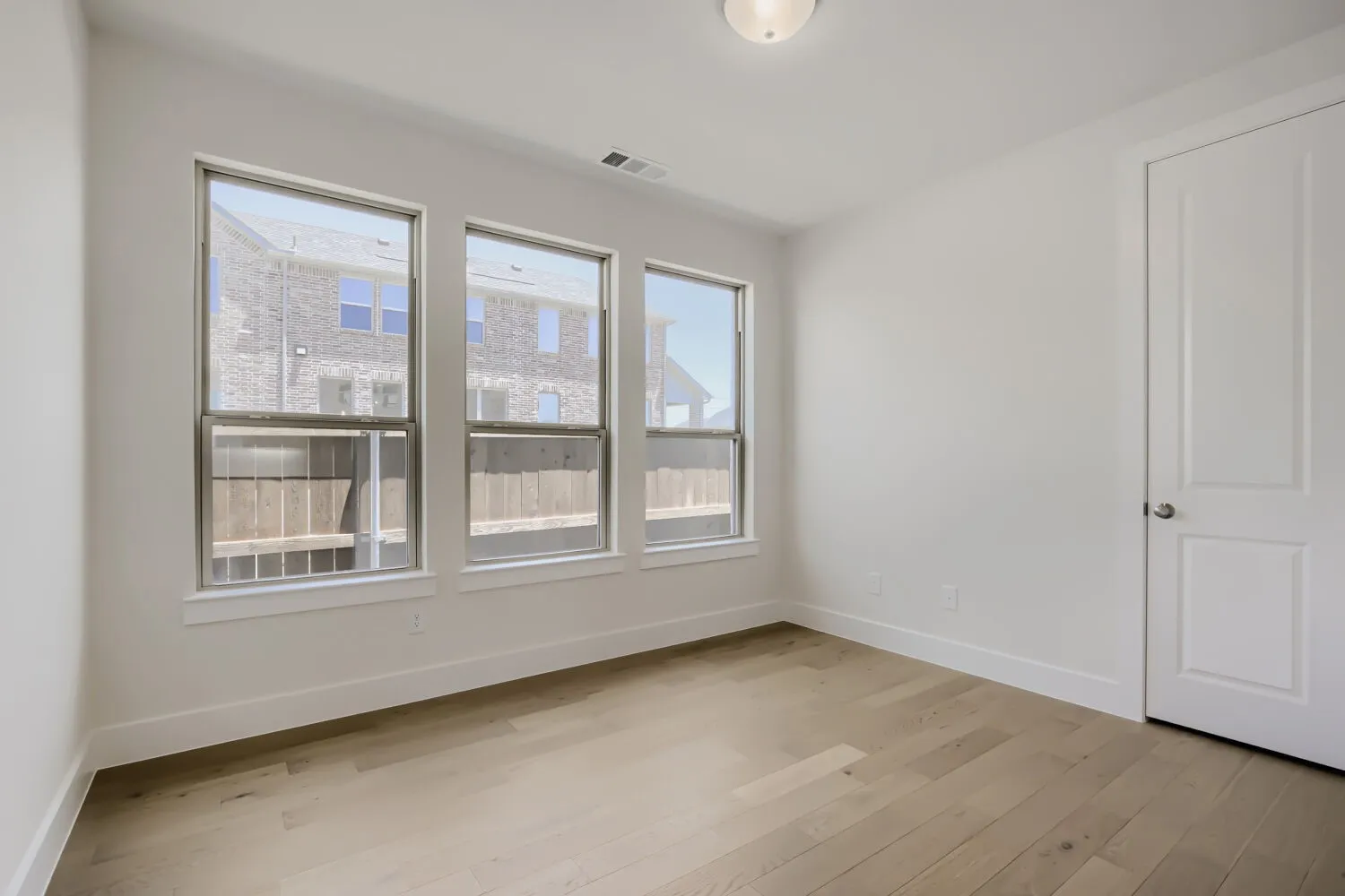 Room featuring three large windows, light wood-finish flooring, white baseboards, a white paneled door, and a flush-mount ceiling light fixture