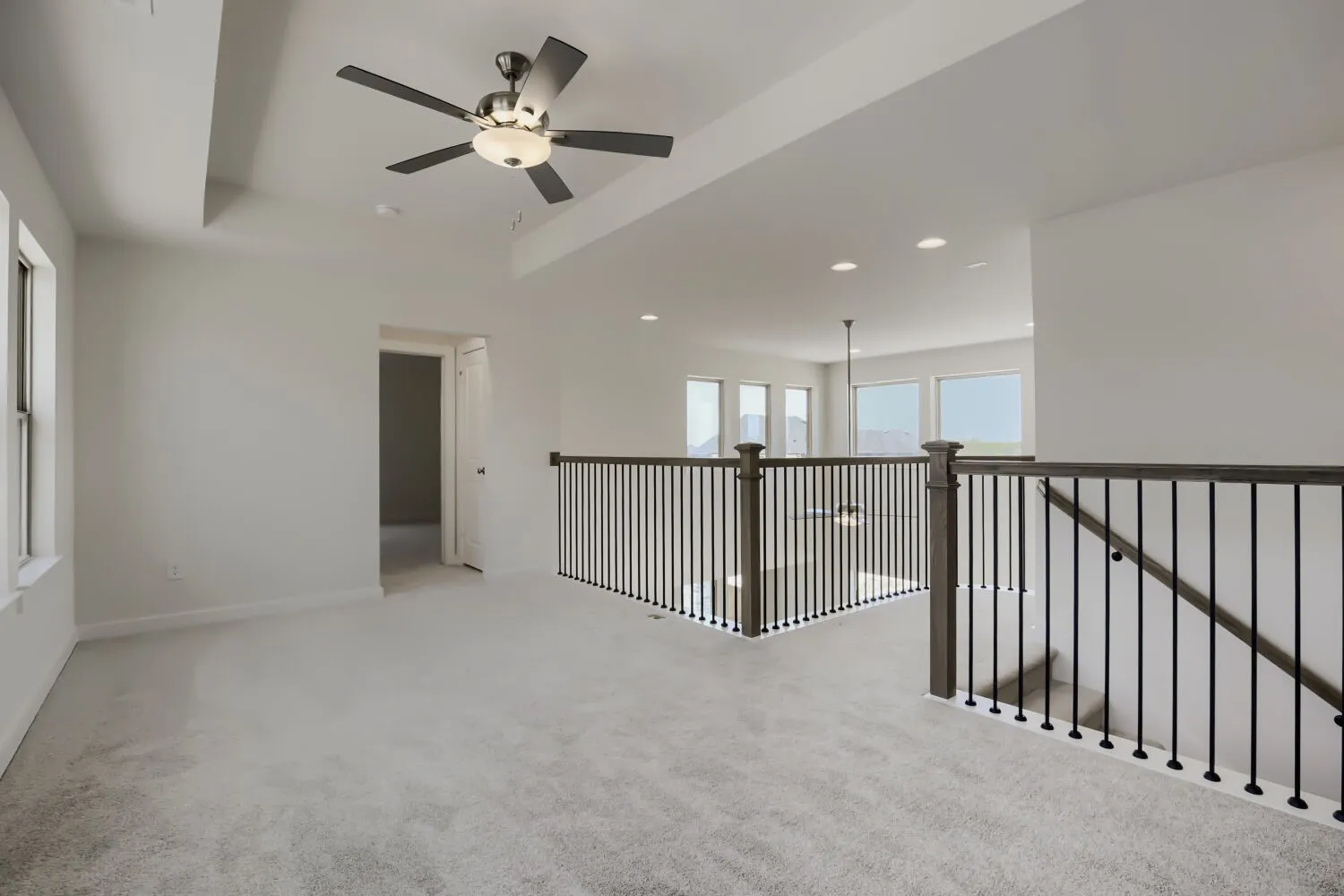 Carpeted loft area featuring a tray ceiling with a modern ceiling fan, large windows, and a dark wood and iron spindle railing overlooking an open space with additional windows