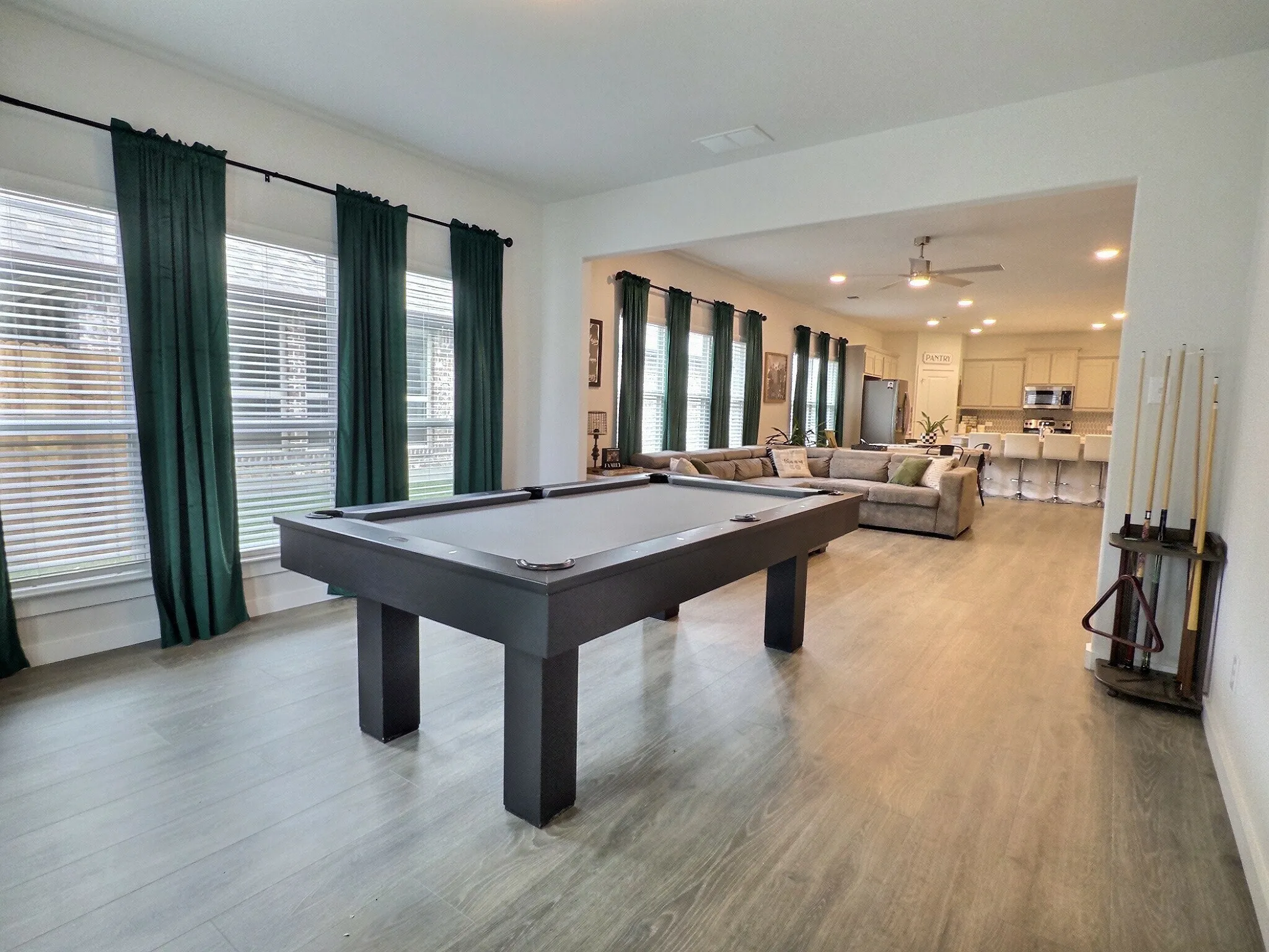 This expansive open-concept room features light-colored flooring, windows with blinds and dark green curtains, and recessed lighting