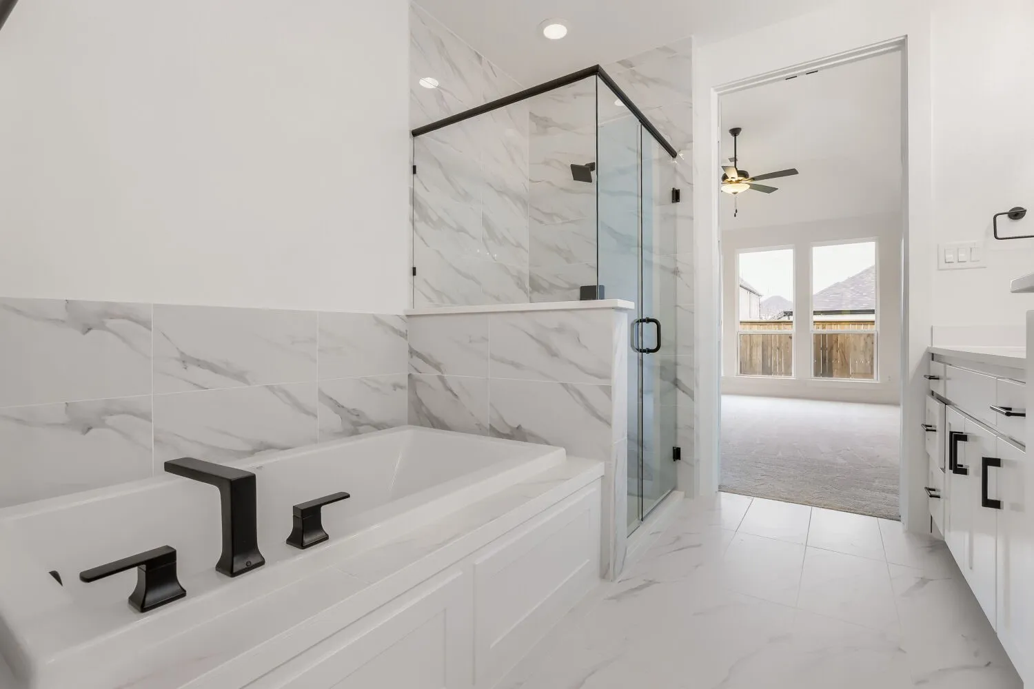 Soaking tub with matte black fixtures, glass-enclosed shower, white cabinetry with black hardware, and marble-look tile flooring