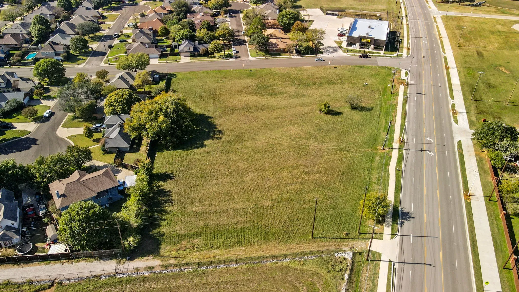 This property features a large, undeveloped grass lot with scattered trees, bordered by an asphalt road and a residential area