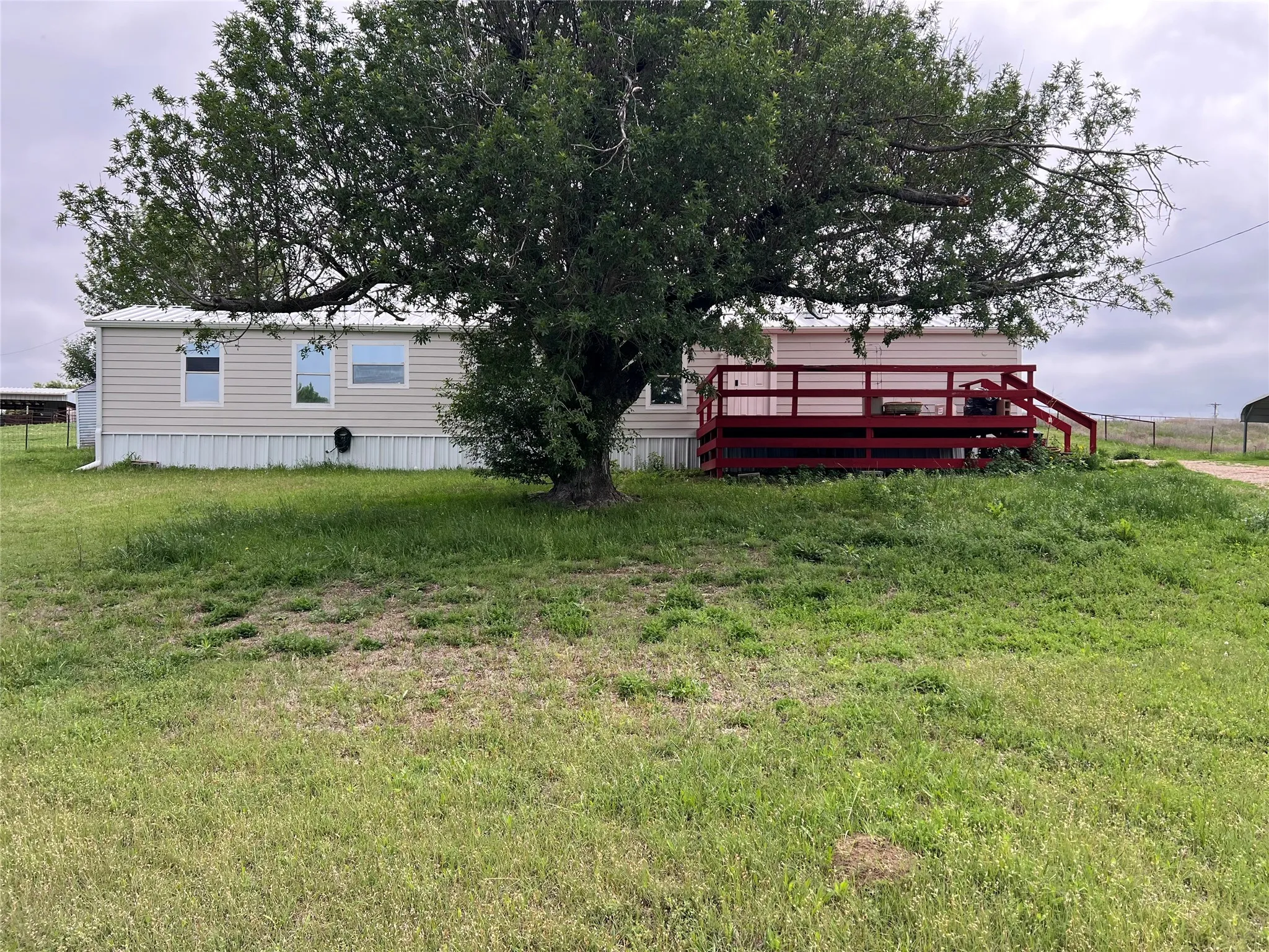 The property features a large yard with a mature tree providing shade, a red-painted deck with railings and steps, and a light-colored exterior with multiple windows