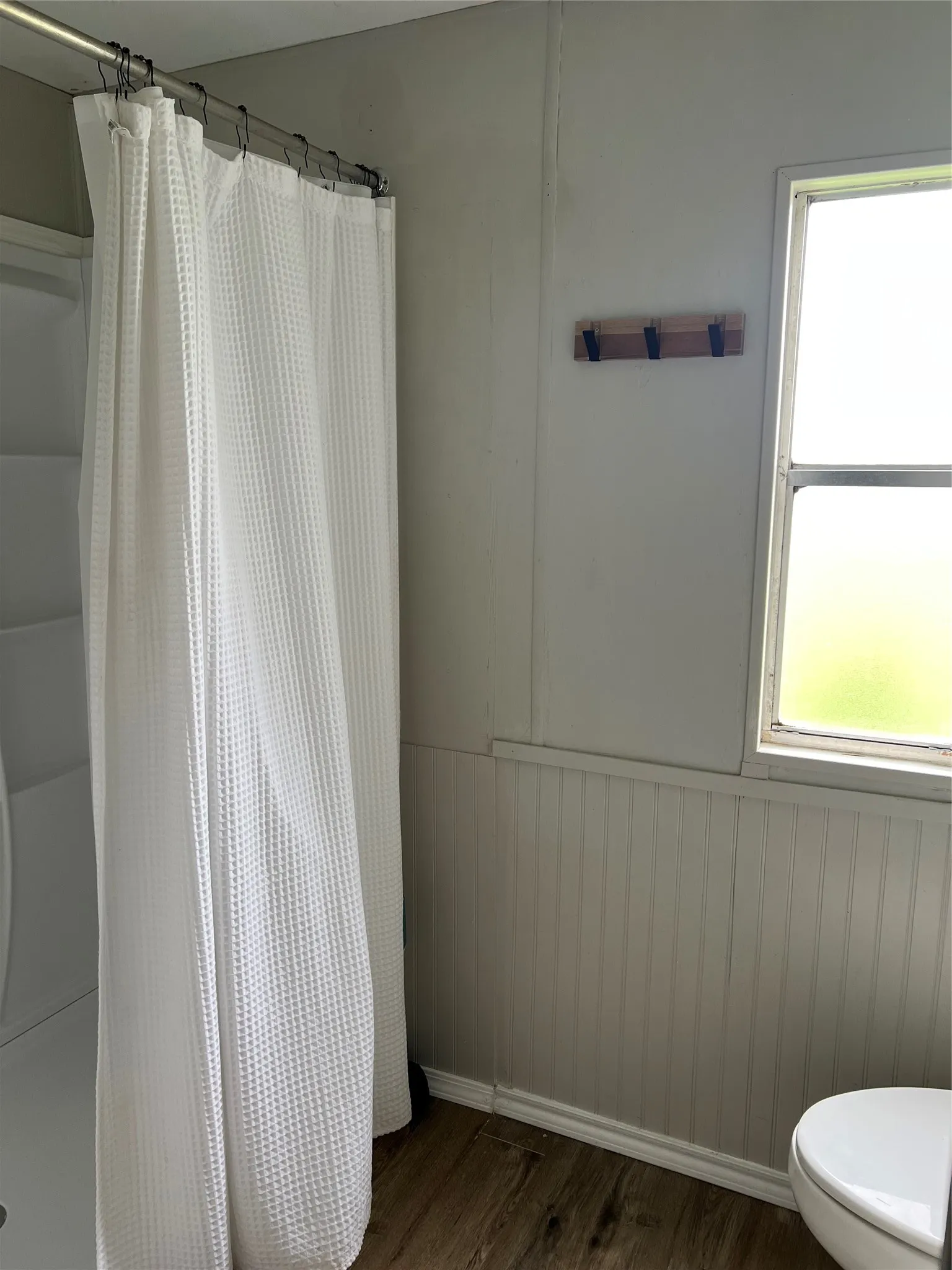 The bathroom features a shower with a white waffle-weave curtain, wood-look flooring, and a window providing natural light