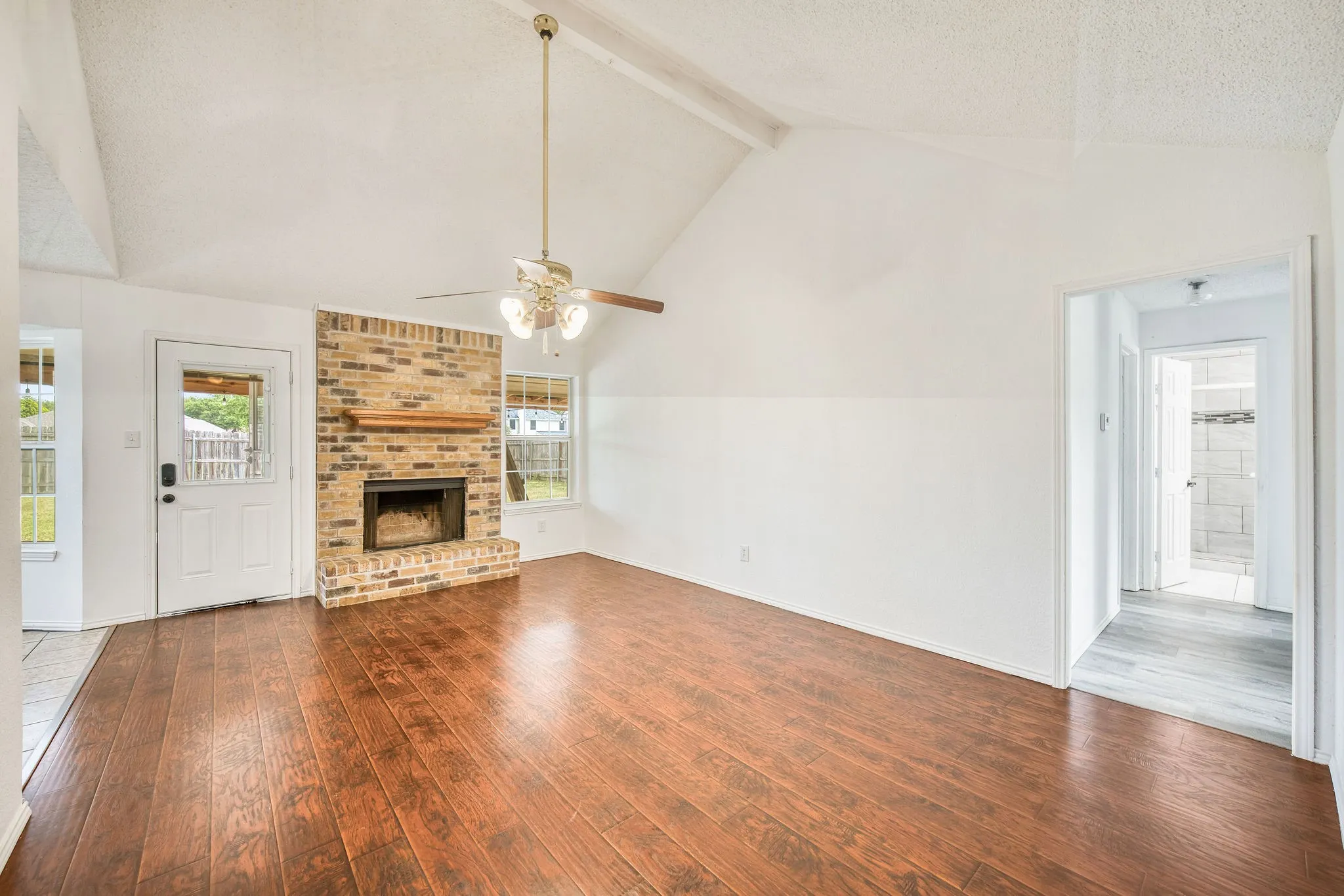Spacious living area with vaulted ceilings, hardwood floors, a cozy brick fireplace with a wood mantel, and a ceiling fan for added comfort