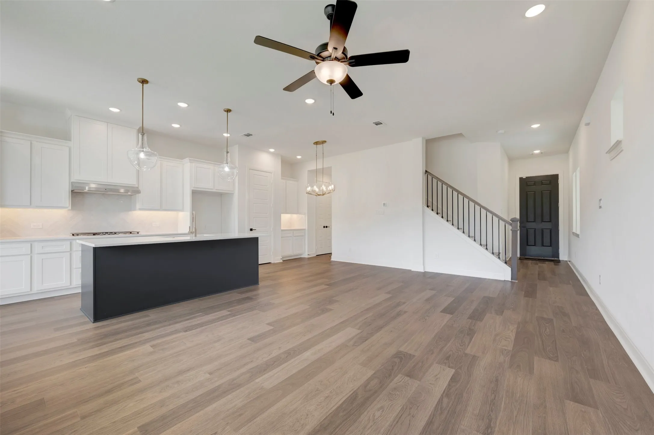 Spacious open-concept layout featuring wood-finish flooring, a kitchen with a dark-toned island and white cabinetry, recessed lighting, and a modern ceiling fan