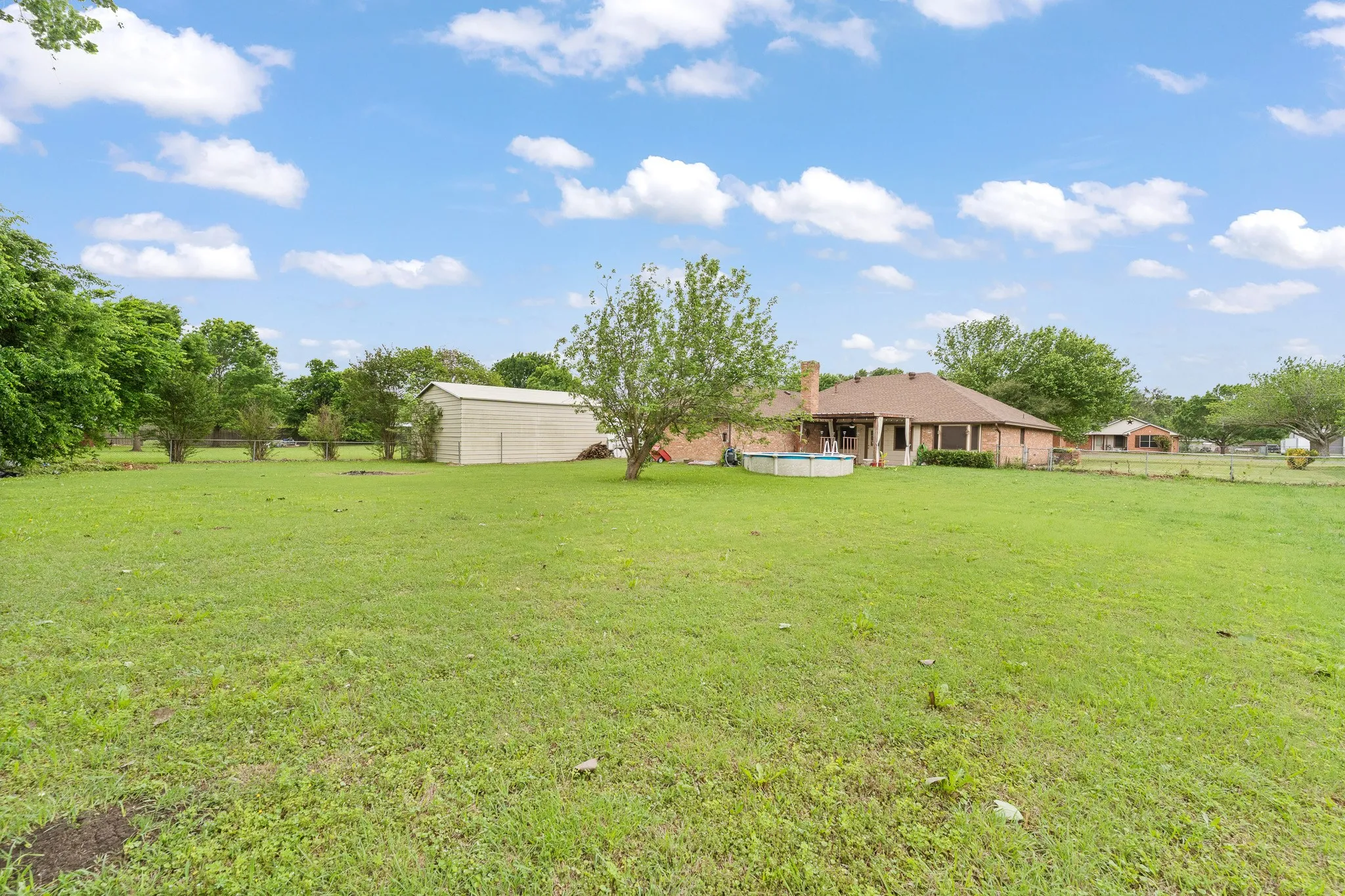 Expansive yard with green lawn, mature trees, a detached storage building, and a brick exterior property with a covered patio