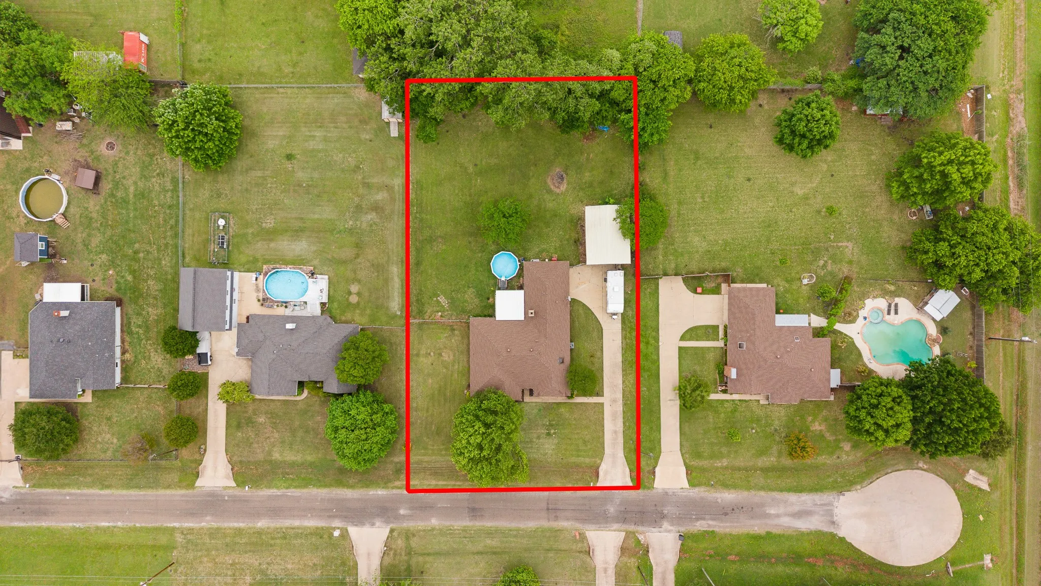 Aerial view of the property, featuring a residential home with a brown roof, a concrete driveway, and a spacious lawn with mature trees