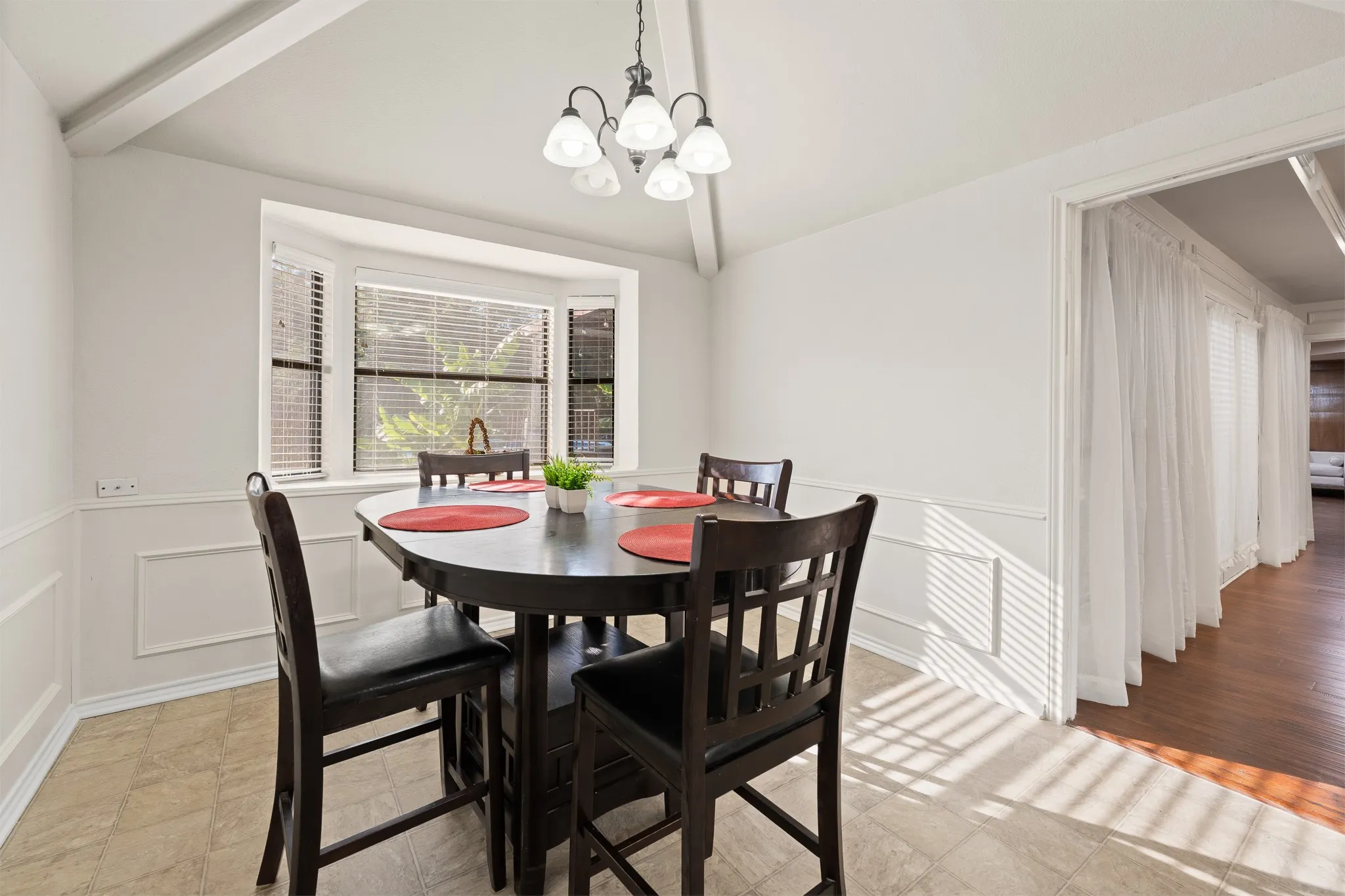 Bay window with blinds, decorative wainscoting, exposed ceiling beams, and overhead chandelier