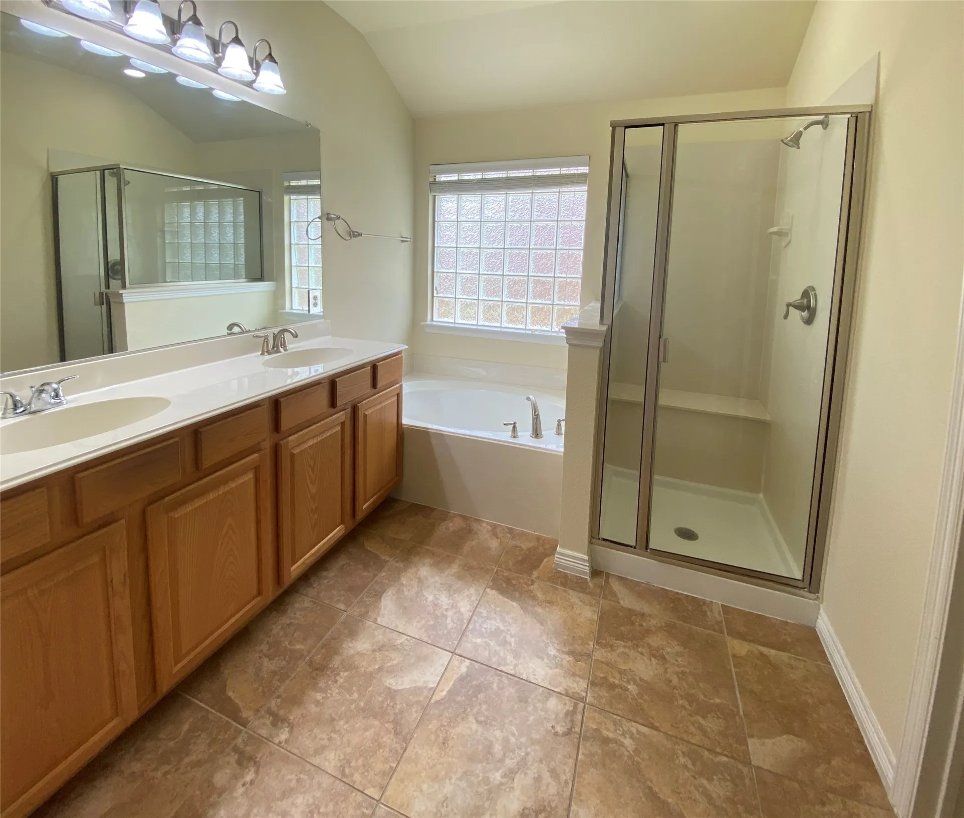 Master Bathroom featuring a dual vanity with light wood cabinetry, a built-in soaking tub, a separate glass-enclosed shower with a bench, privacy glass block window, and large format floor tiling
