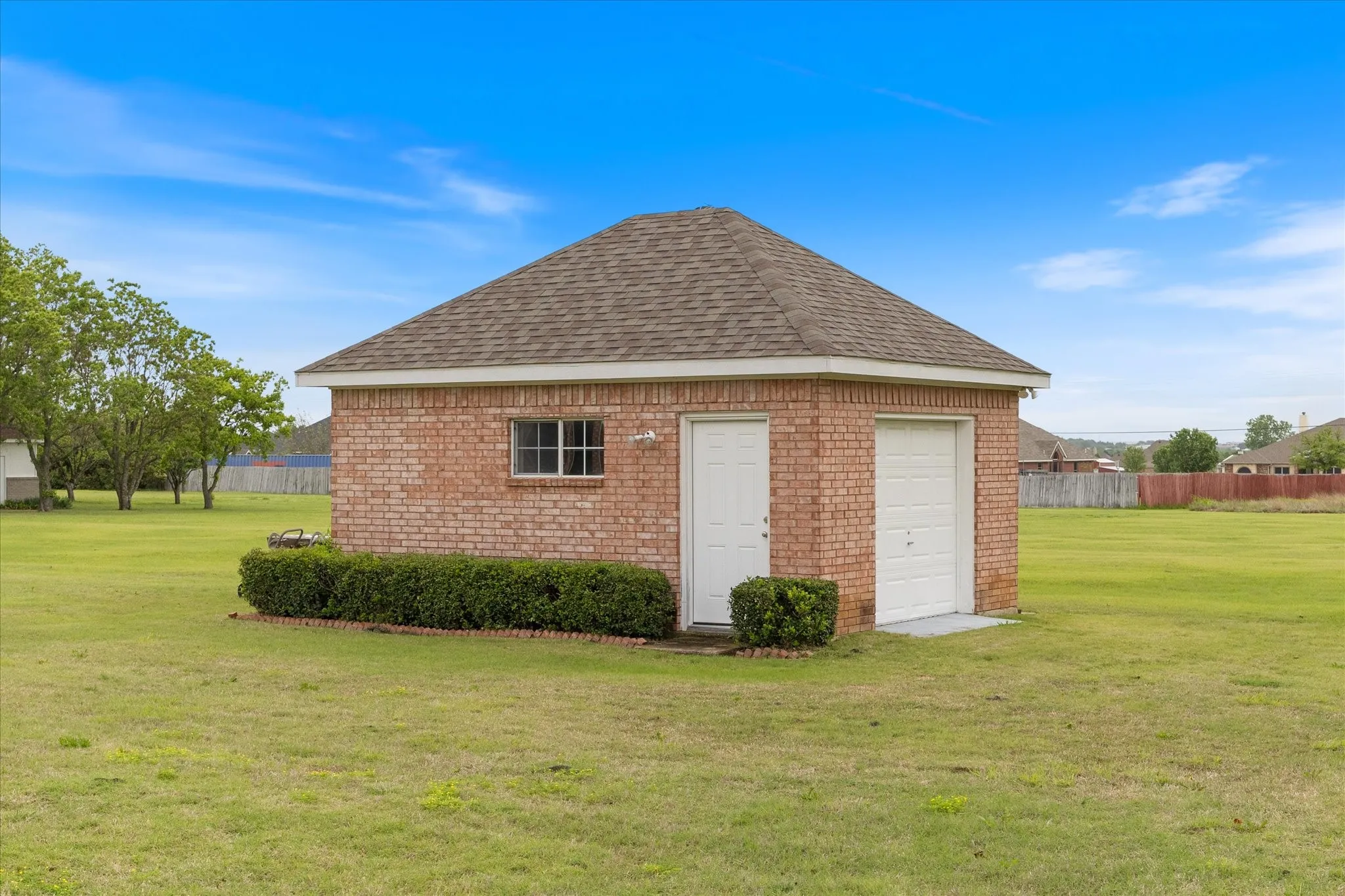 Detached brick building with a shingled roof, featuring a single window, a side entry door, and two garage doors, all set within a expansive grassy area