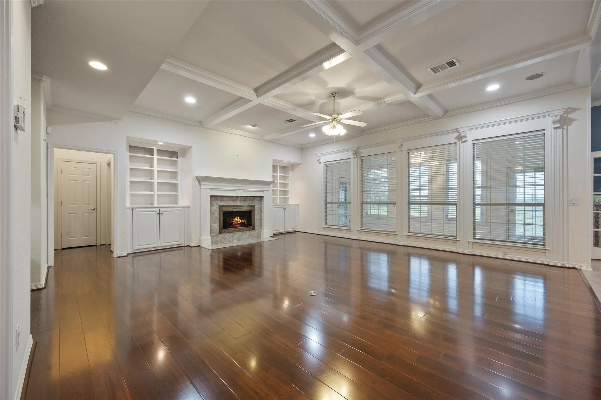 Living area featuring shining wood floors, a coffered ceiling with a ceiling fan, a fireplace with a stone surround and mantel, and built-in shelving and cabinetry