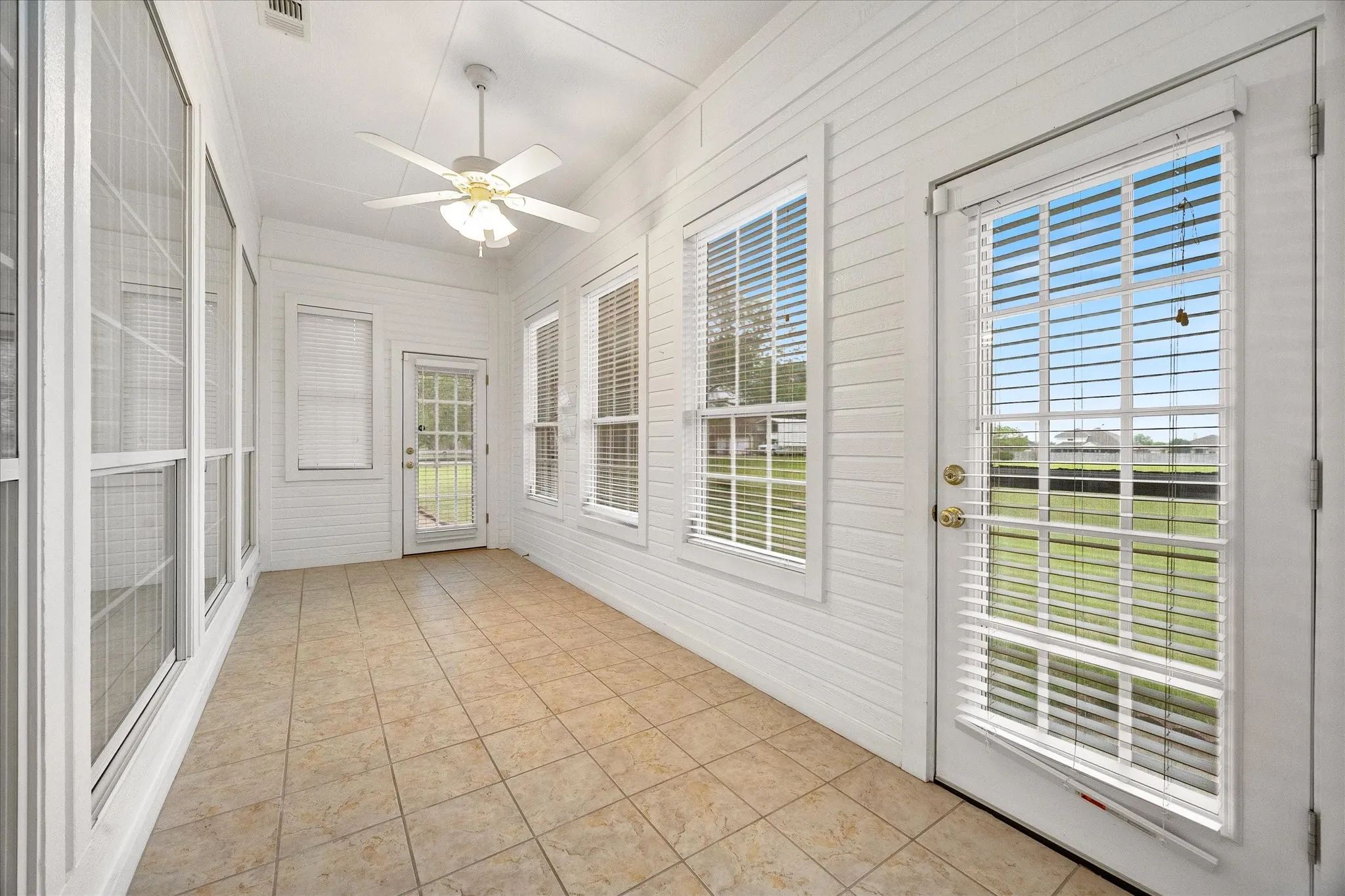 The sunroom features tiled flooring, white paneling, a ceiling fan, and abundant natural light from numerous windows and glass-paneled doors