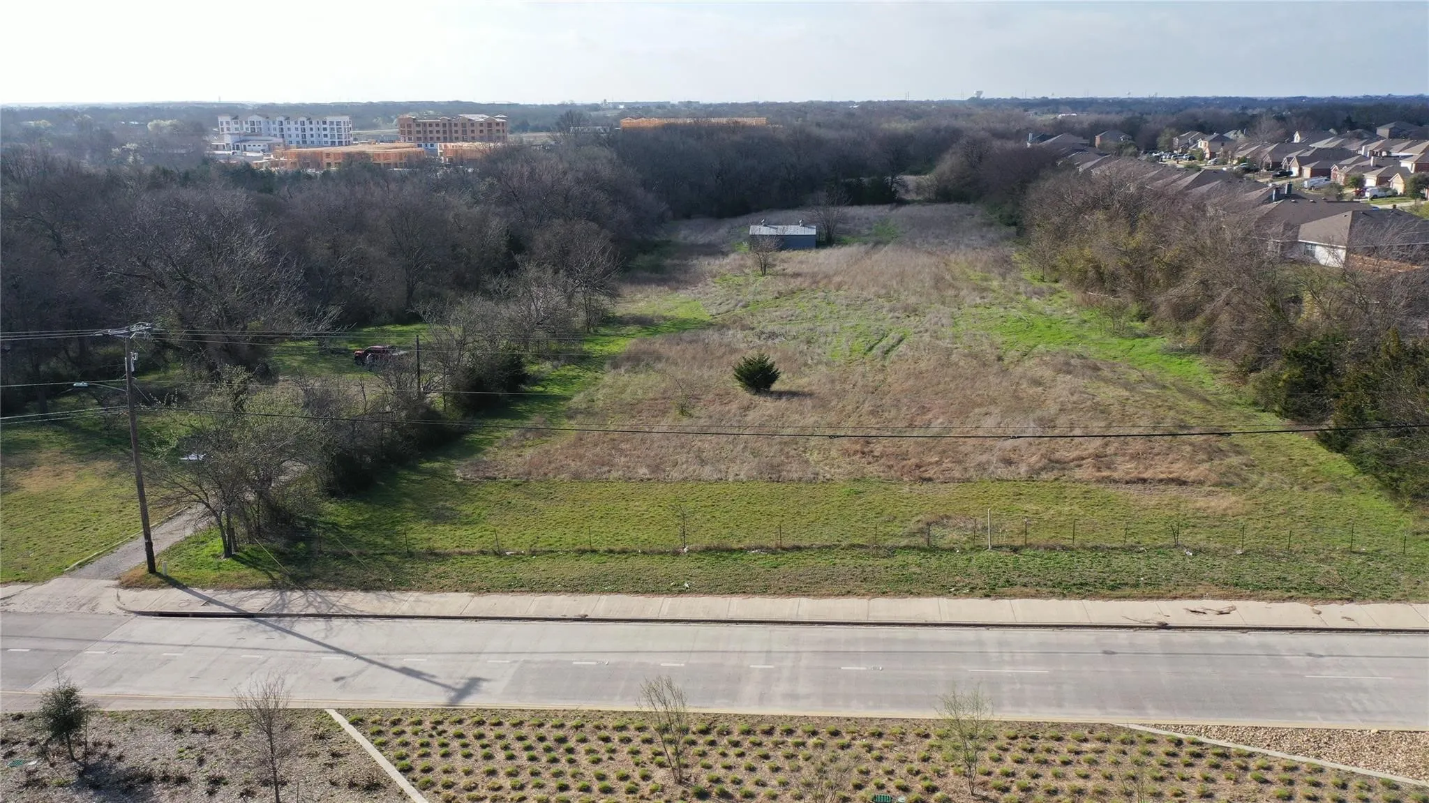 Expansive undeveloped lot with a visible road frontage, surrounded by trees and residential properties in the distance