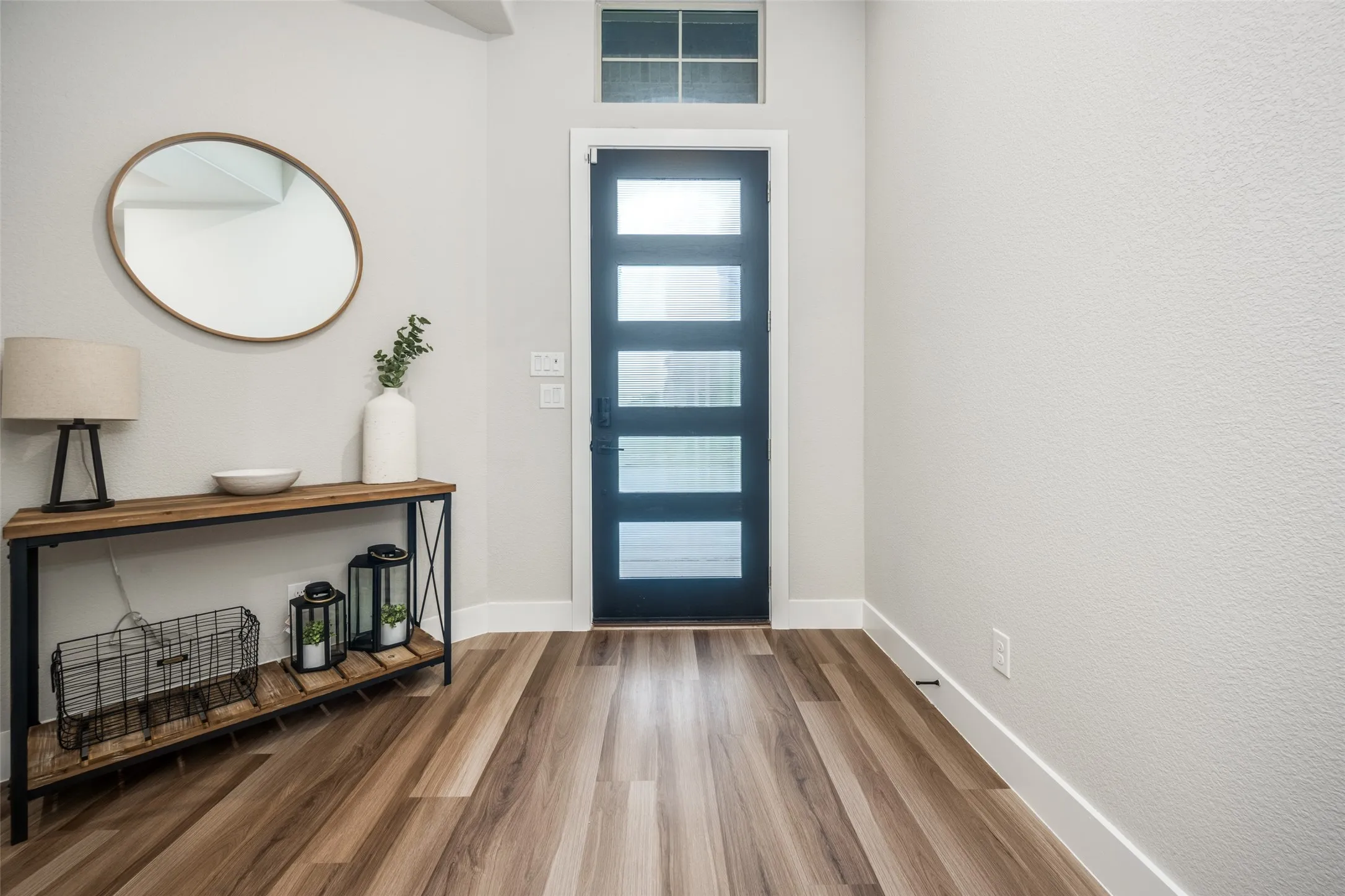Entryway featuring wood-finish flooring, a contemporary black door with frosted glass panels, an overhead transom window, and crisp white baseboards. Open, clean, and bright!