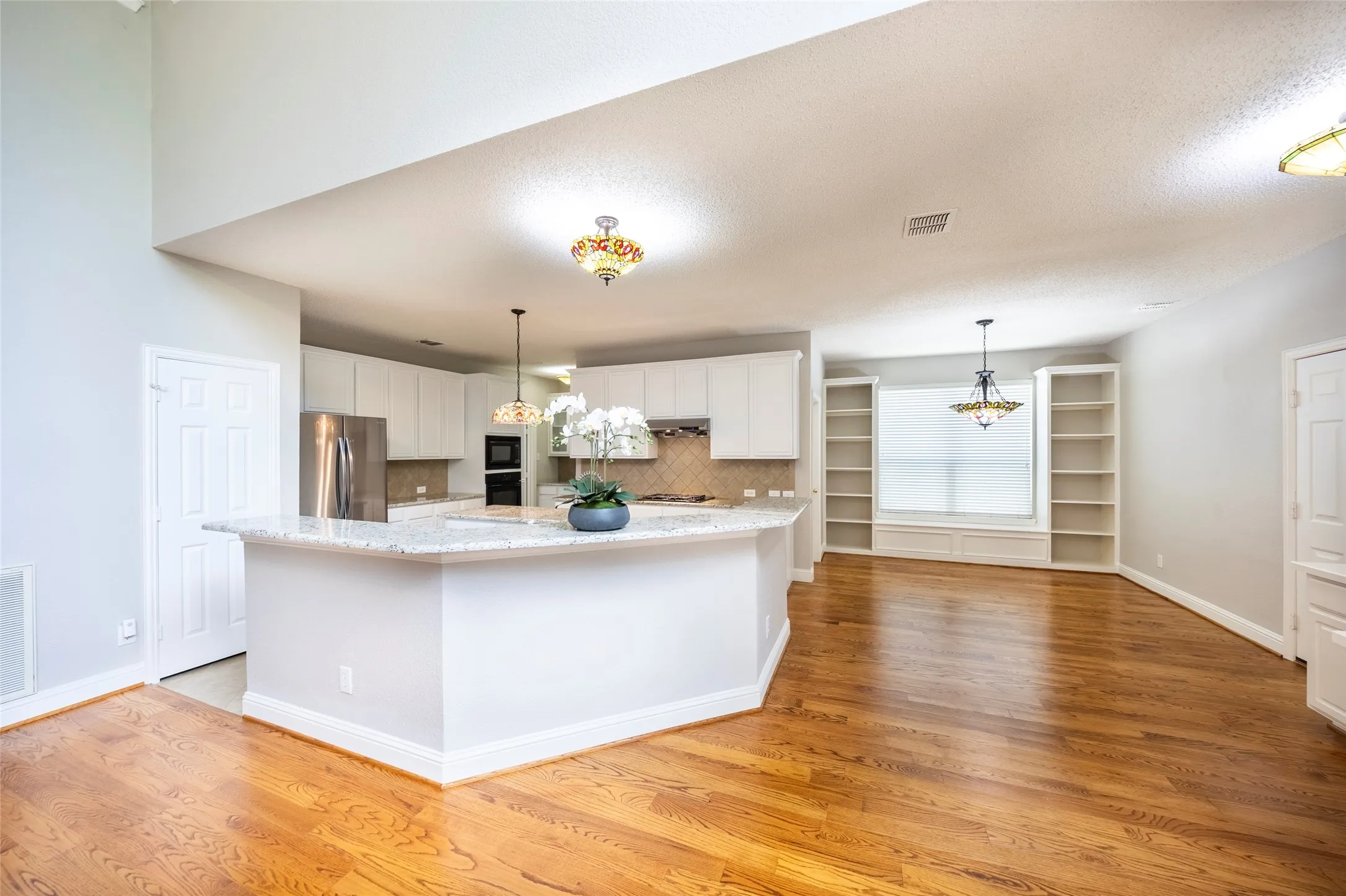 Expansive open-concept living area featuring hardwood flooring, a kitchen island with a granite countertop, white cabinetry, and built-in shelving with a window seat