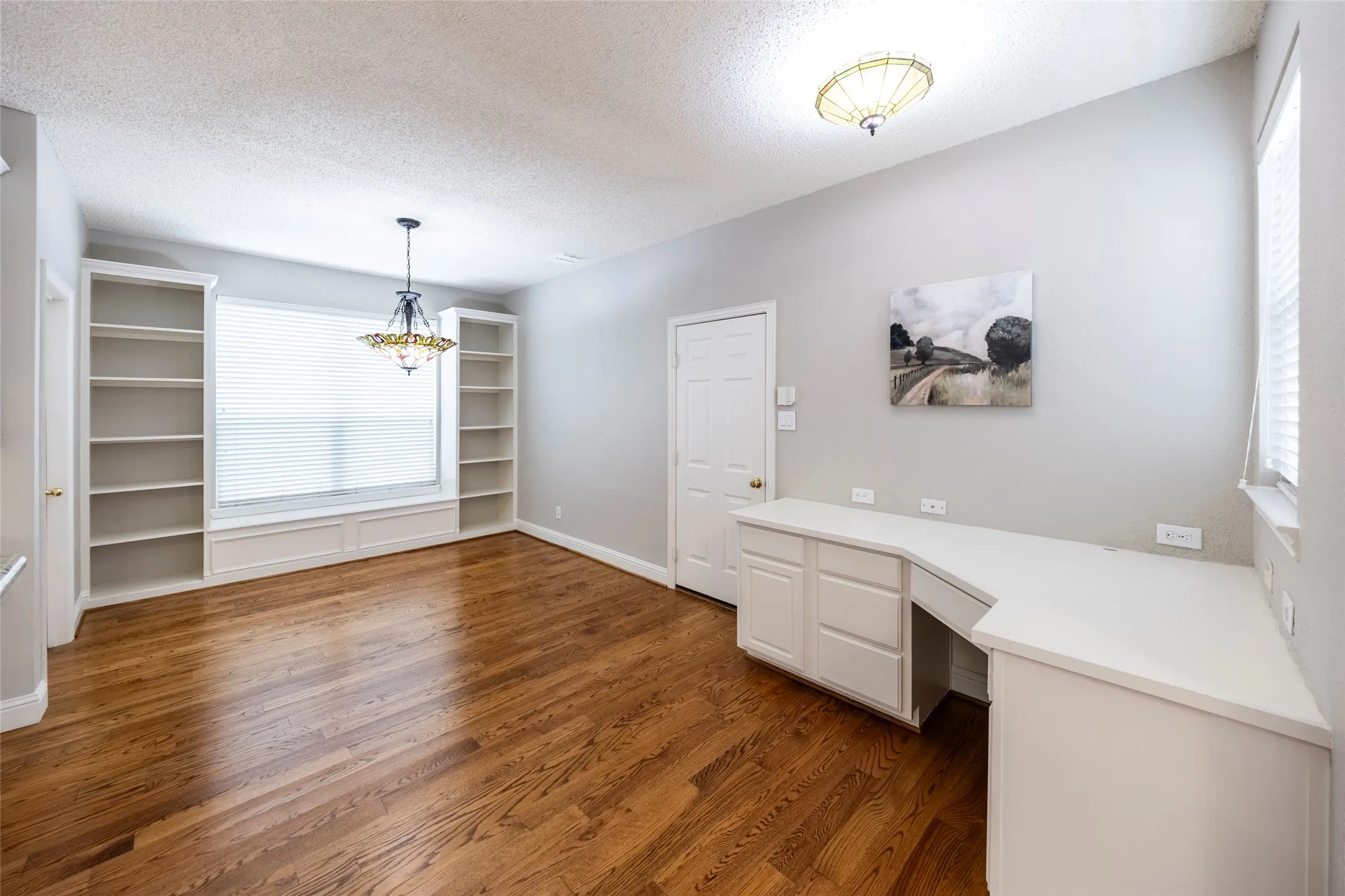 The room features warm hardwood floors, a large window with white blinds, and built-in white shelving units