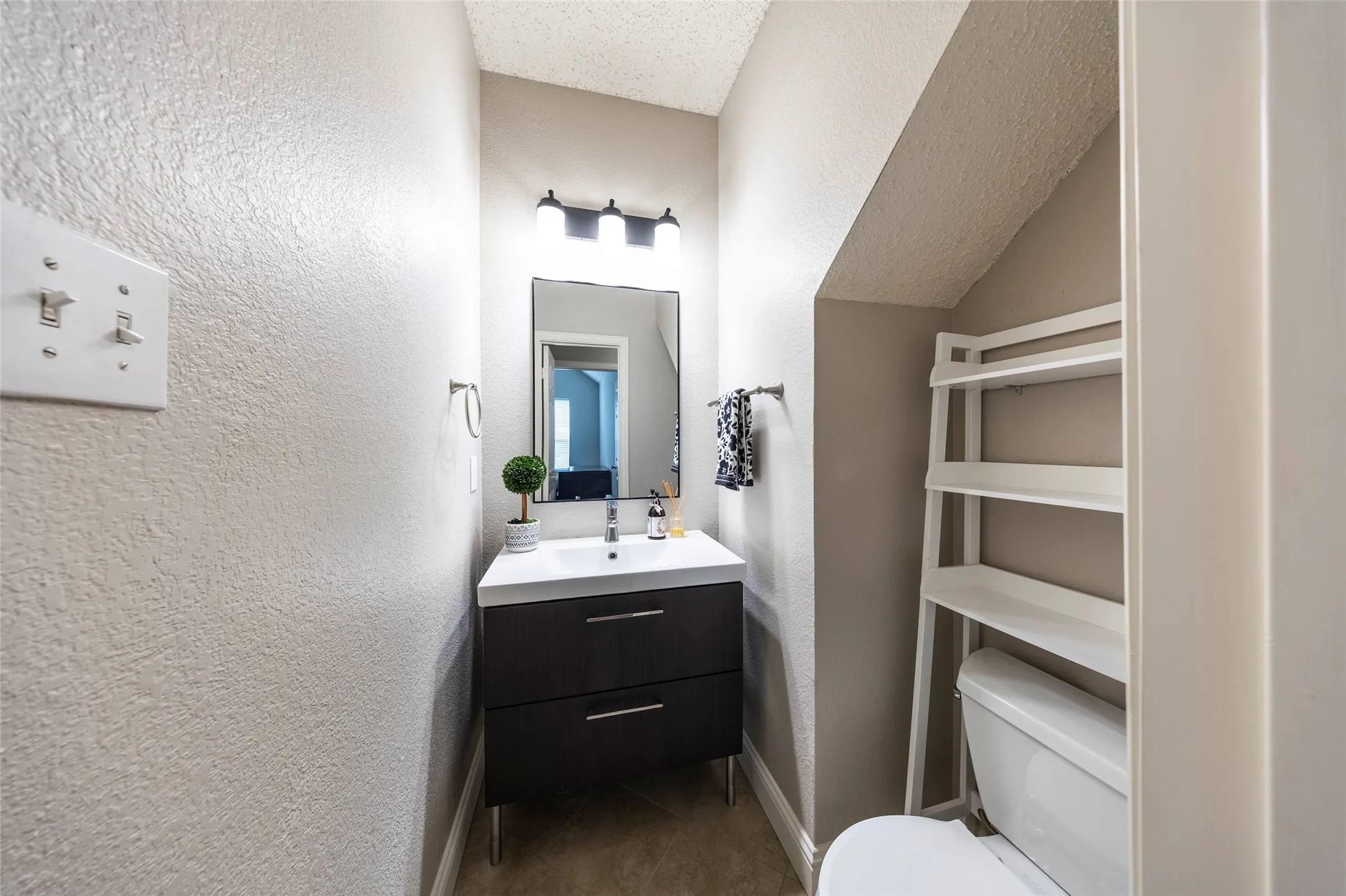 This bathroom features a contemporary vanity with a white countertop and dark cabinetry, complemented by a wall-mounted mirror and overhead lighting