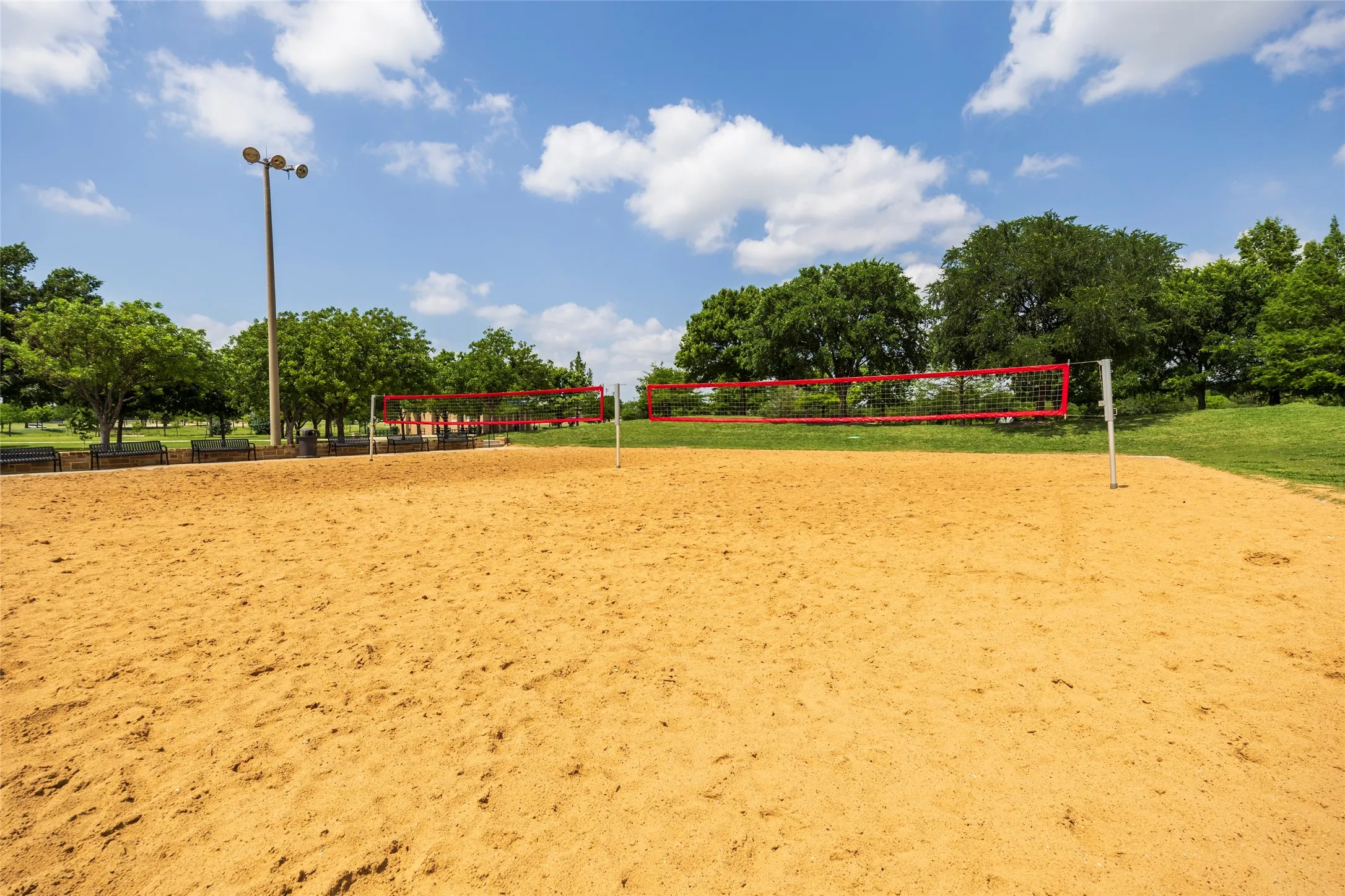 This community amenity features a sand volleyball court with a red net, surrounded by green grass and mature trees