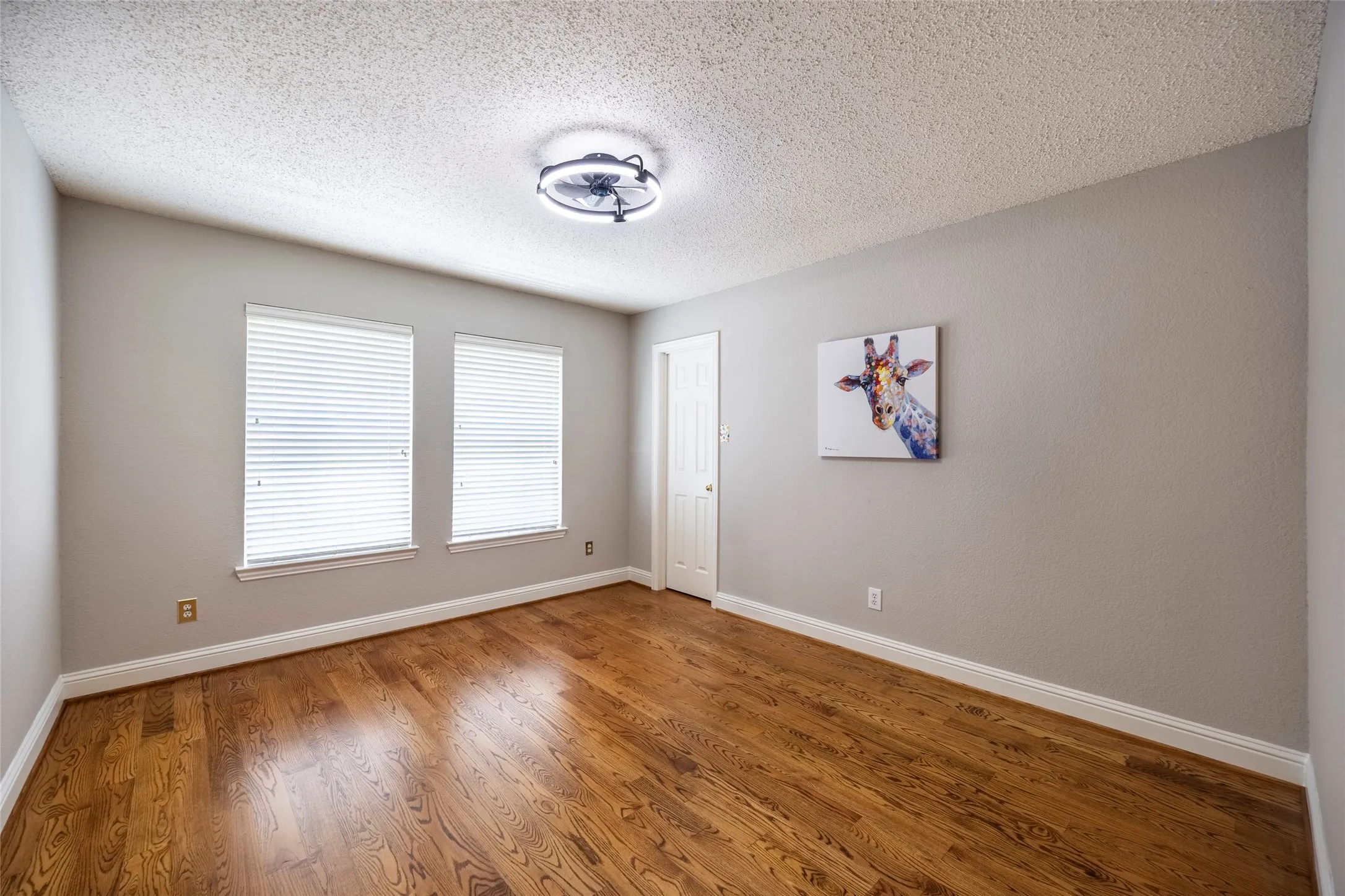 This room features hardwood floors, light gray walls, two windows with blinds, a white door, and a ceiling light fixture